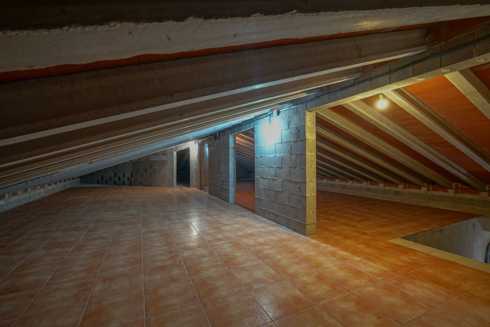 Grenier de rangement avec carrelage au sol, poutres de plafond en béton apparentes et cloisons en parpaings avec éclairage intégré.