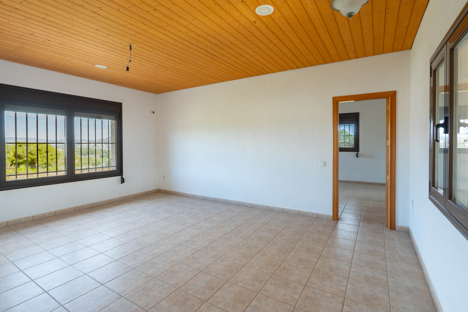 Pièce avec plafond en lambris de bois, carrelage beige et fenêtres à cadres sombres munies de barreaux de sécurité offrant une vue dégagée.