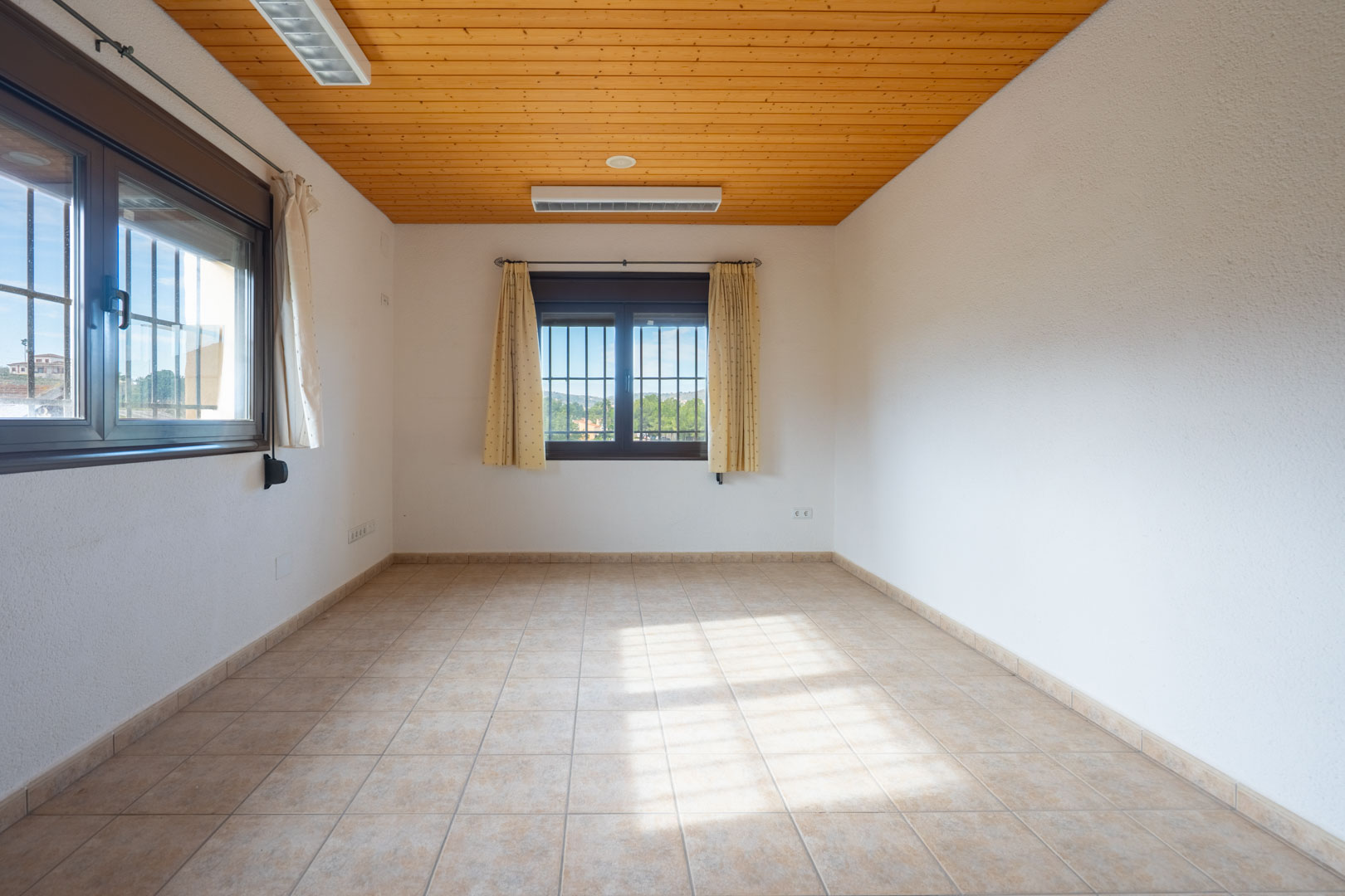 Pièce lumineuse avec plafond en bois, sol en carrelage beige et deux fenêtres sécurisées par des grilles et des rideaux clairs.