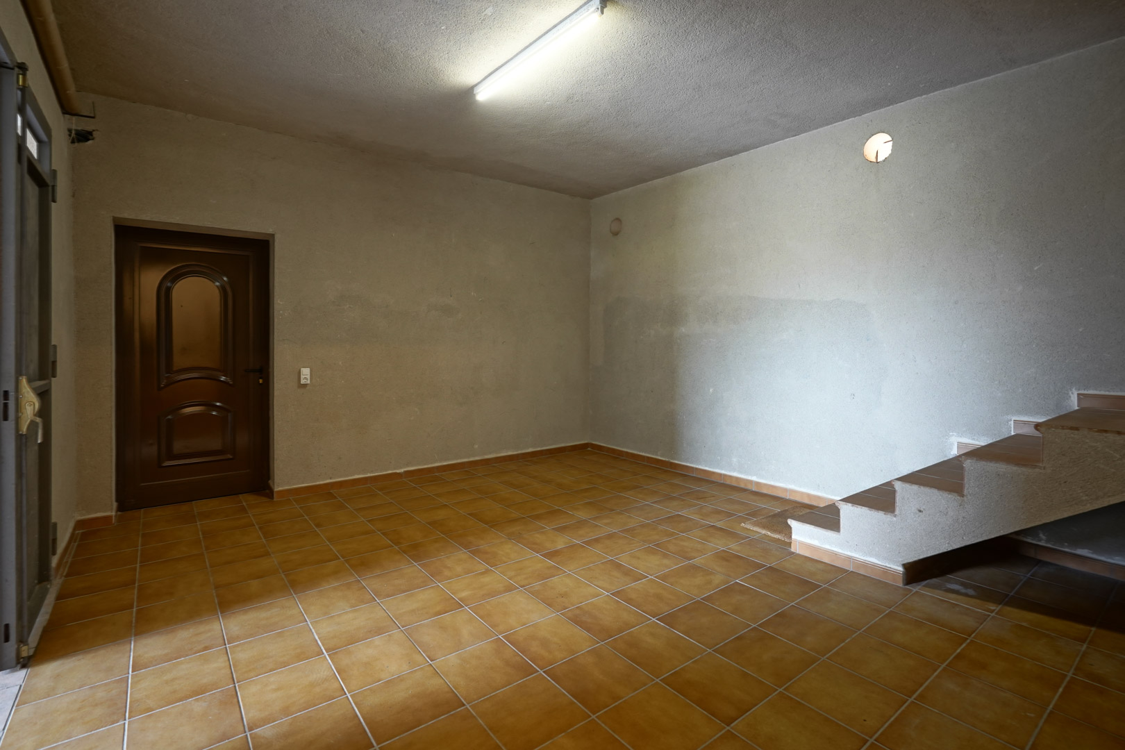 Pièce polyvalente avec sol en grès cérame, murs à l'aspect brut et escalier carrelé menant aux étages supérieurs.