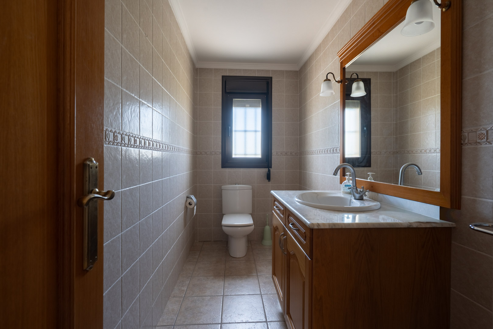 Salle de bain avec meuble en bois, plan en marbre et grand miroir. Les murs sont revêtus de carreaux beiges avec une bordure décorative et une fenêtre.