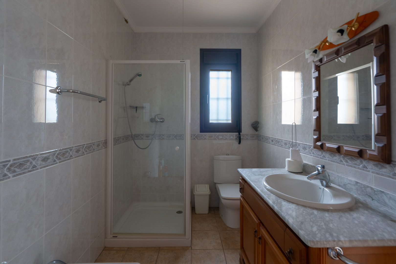 Salle de bain avec plan de toilette en marbre, meuble en bois et cabine de douche vitrée. Murs carrelés avec frise décorative et fenêtre à verre dépoli.