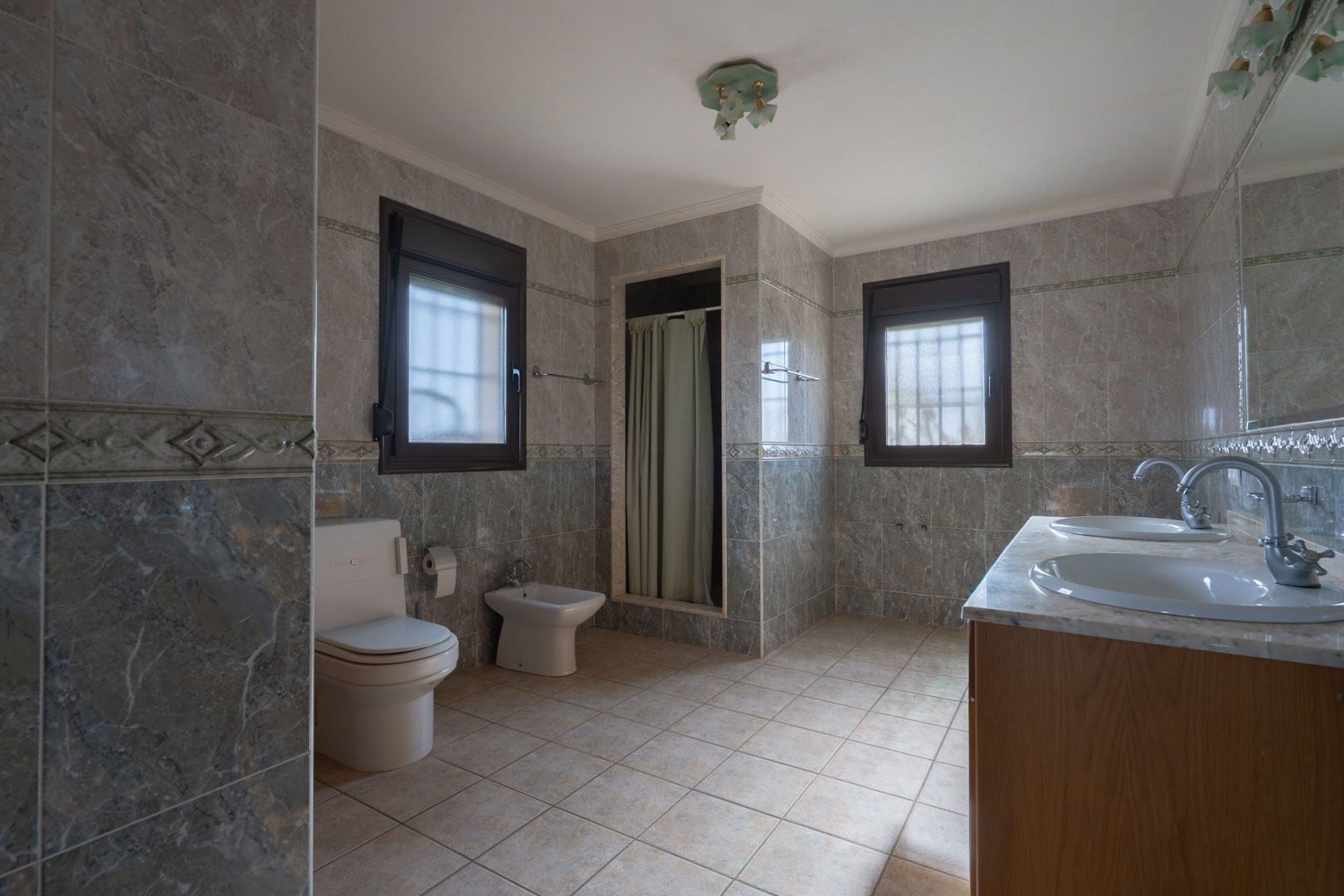 Salle de bain spacieuse avec double vasque en marbre, carrelage mural gris à motifs et deux fenêtres offrant une lumière naturelle diffuse.