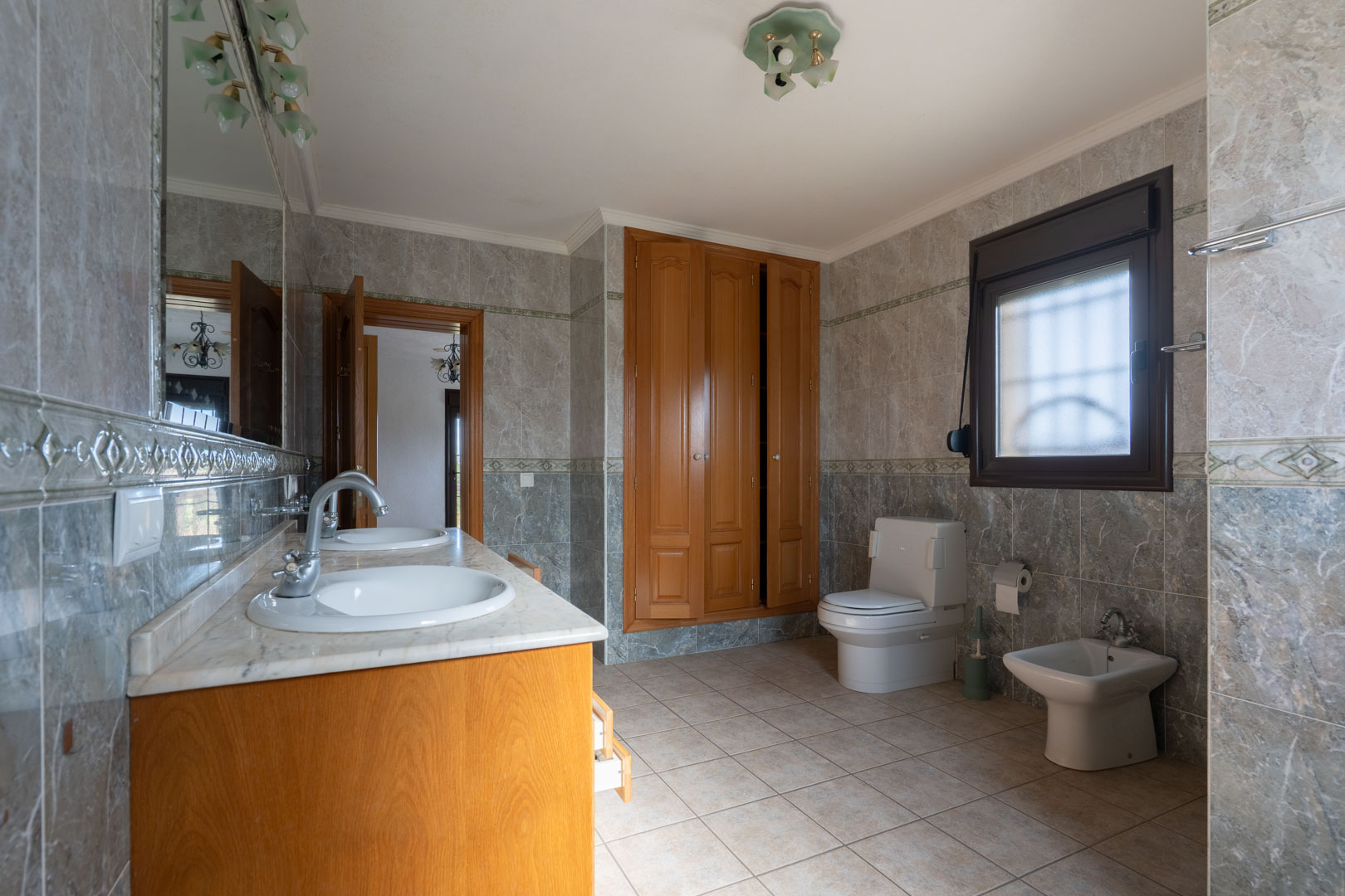 Salle de bain spacieuse avec double vasque en marbre, placards intégrés en bois et murs entièrement carrelés avec frises décoratives. Comprend WC et bidet.