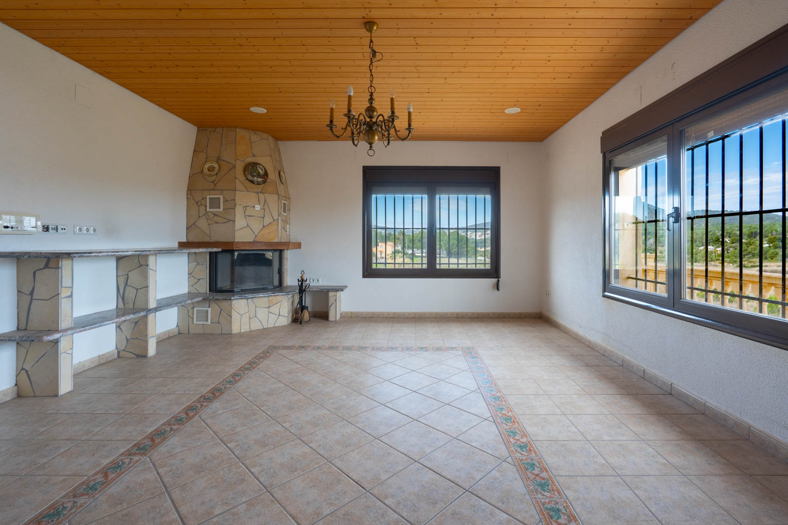 Salon avec plafond en lambris, cheminée revêtue de pierre naturelle et sol en carrelage avec bordure décorative. Les fenêtres offrent une vue dégagée.