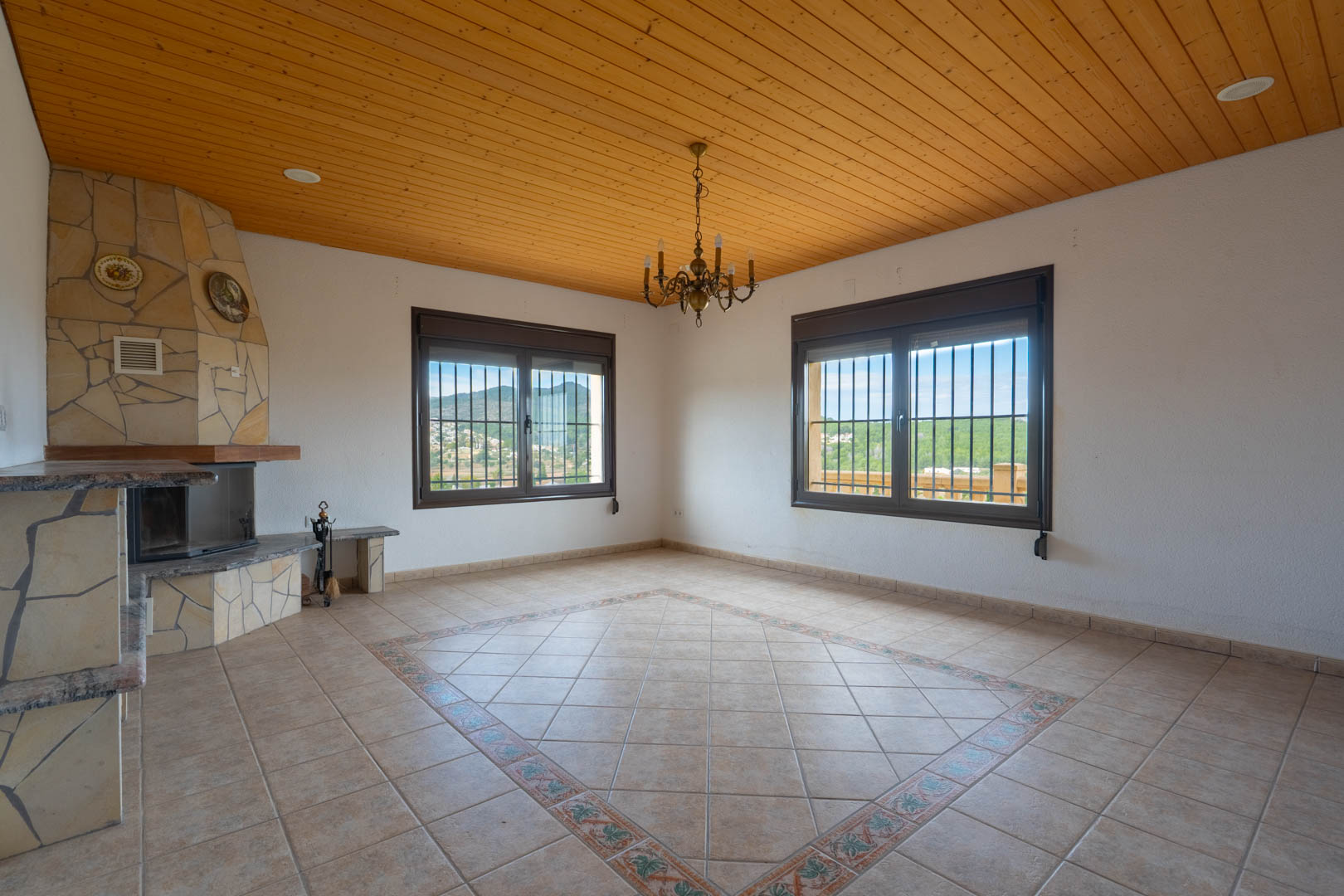 Salon spacieux avec plafond en bois, cheminée d'angle en pierre et sol carrelé avec motif décoratif. Les fenêtres offrent une vue sur le paysage vallonné.
