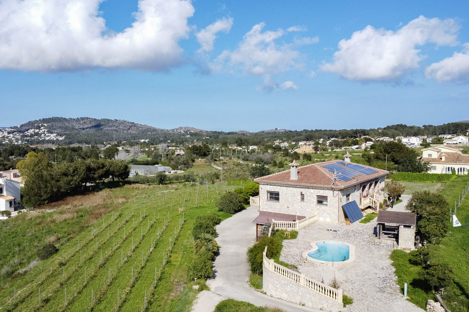 Vue aérienne d'une maison de campagne en pierre avec panneaux solaires, piscine en forme de haricot sur terrasse pavée et vignobles adjacents.