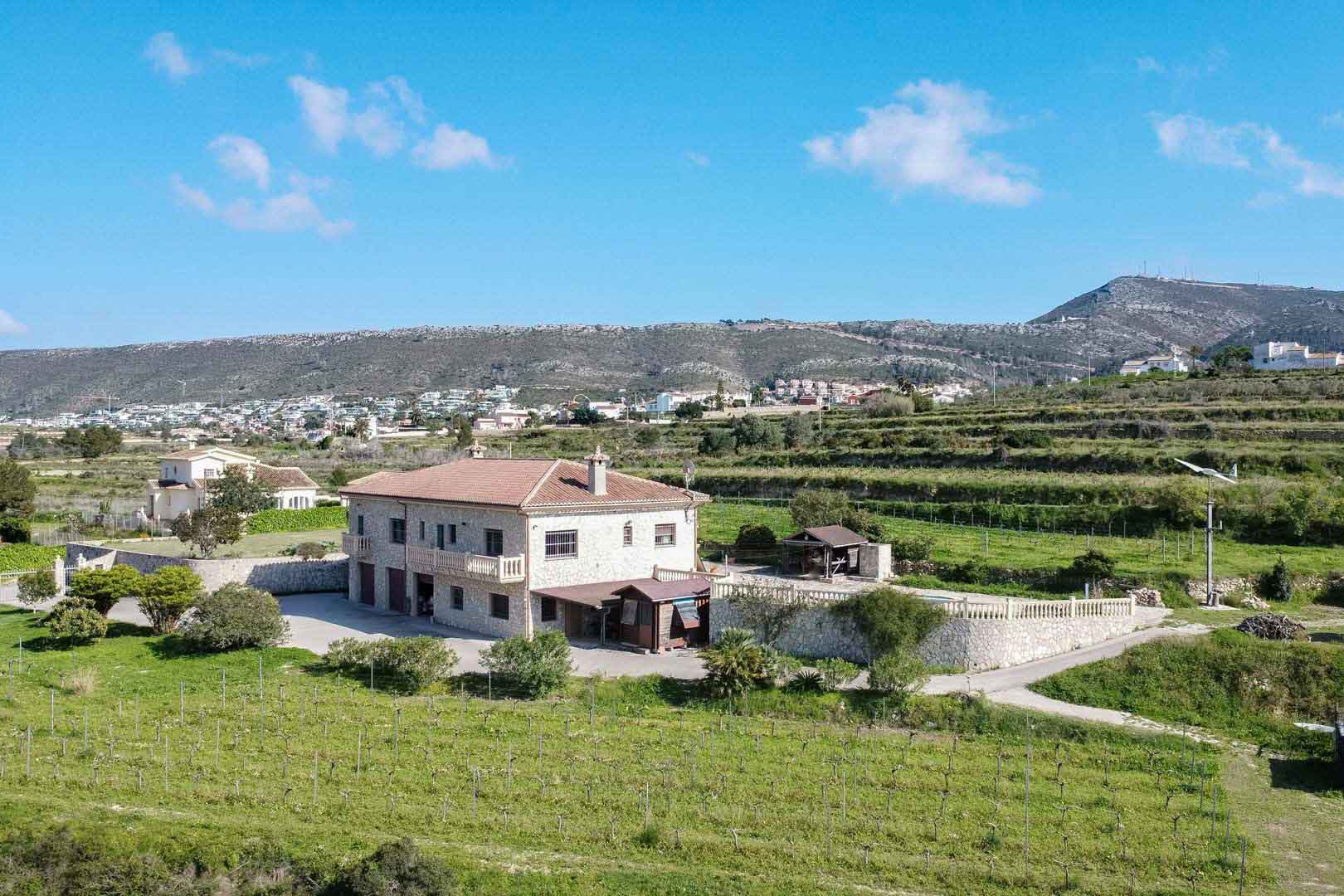 Vue aérienne d'une maison de campagne en pierre avec toit en tuiles, entourée de vignes et de champs verdoyants avec un panorama sur les collines environnantes.