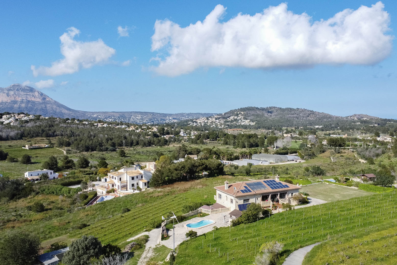 Vue aérienne d'une villa en pierre avec panneaux solaires, piscine privée et éolienne, entourée de vignobles verdoyants et d'un paysage montagneux.