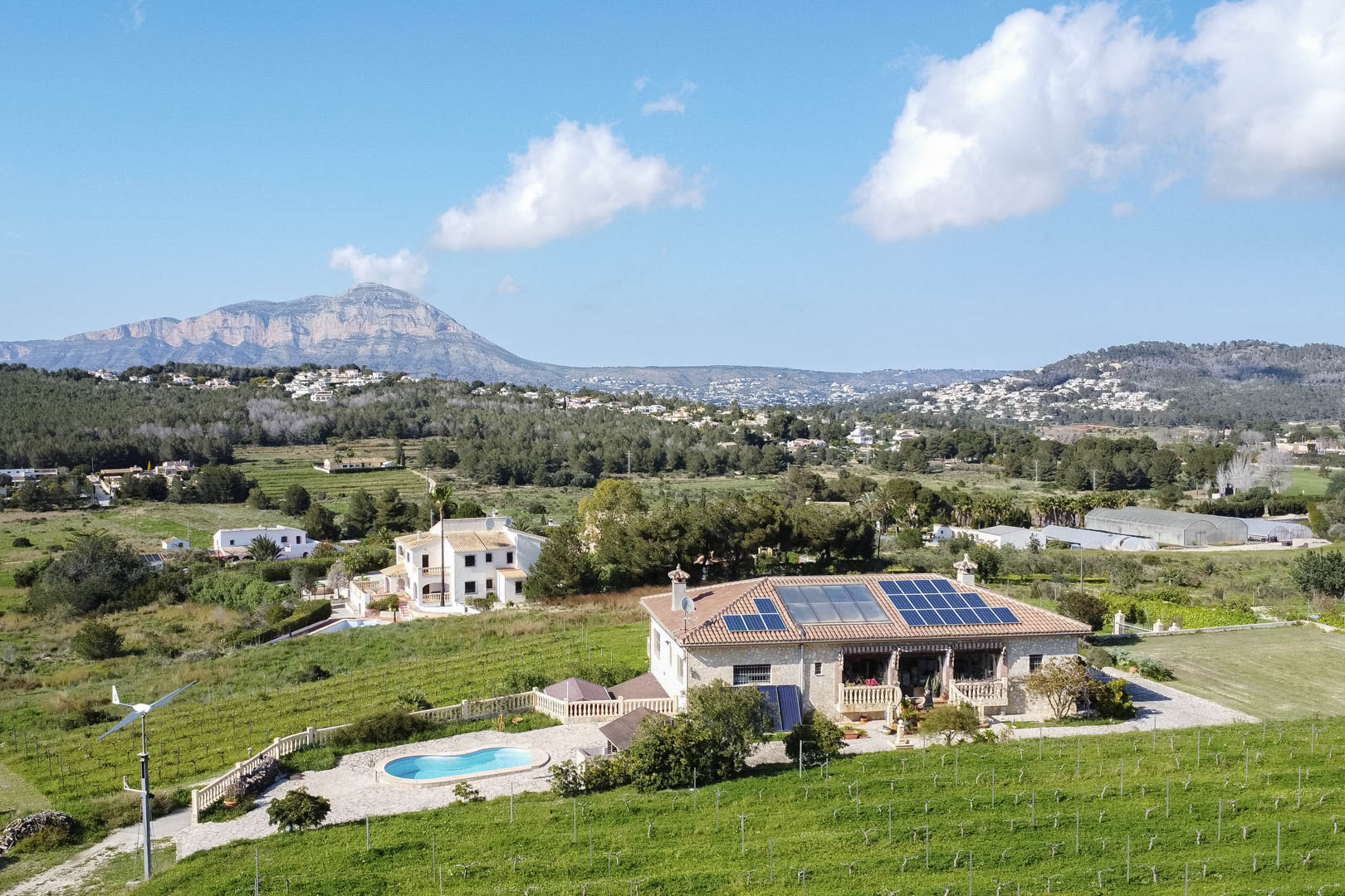 Vue aérienne d'une villa en pierre équipée de panneaux solaires et d'une éolienne, avec piscine privée et vue dégagée sur la chaîne de montagnes.