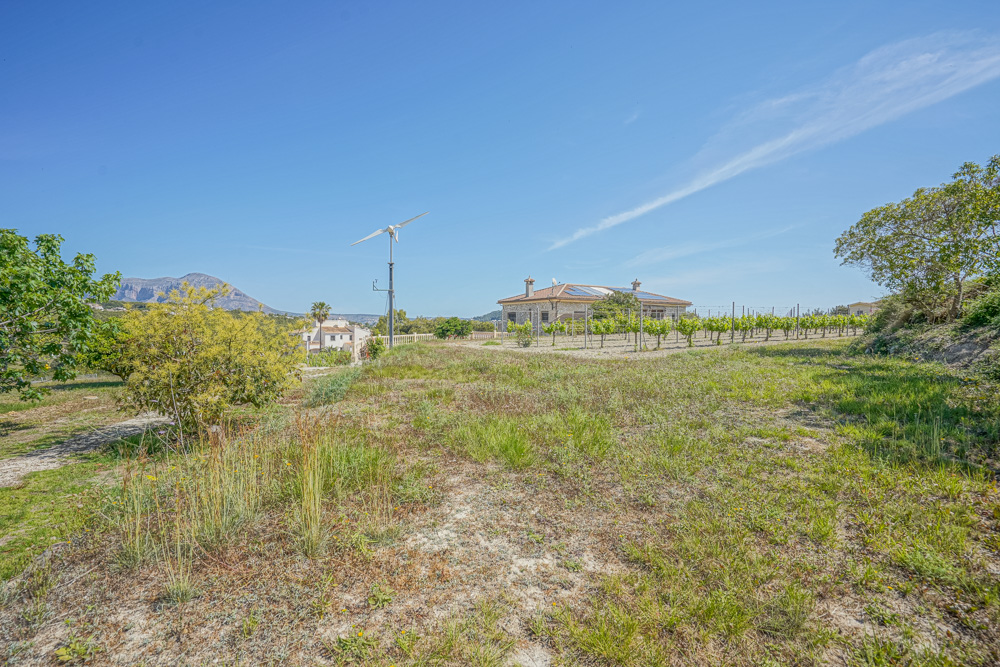 Vue extérieure d'une villa en pierre avec panneaux solaires et éolienne, comprenant un vignoble privé et des vues sur les montagnes environnantes.