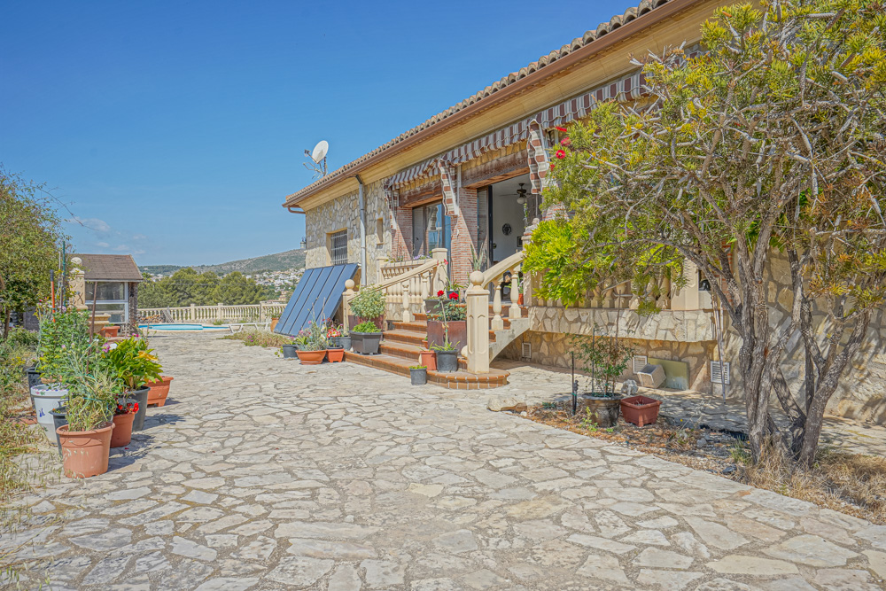 Vue extérieure montrant un patio en pierre, une façade en maçonnerie et des panneaux solaires. Des escaliers mènent à une terrasse couverte à balustres.