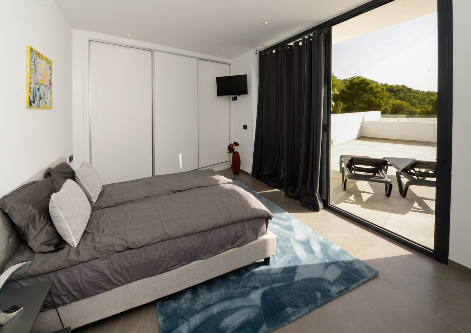 Chambre moderne avec placards intégrés blancs, offrant un accès direct à une grande terrasse avec transats et vue sur les collines verdoyantes.