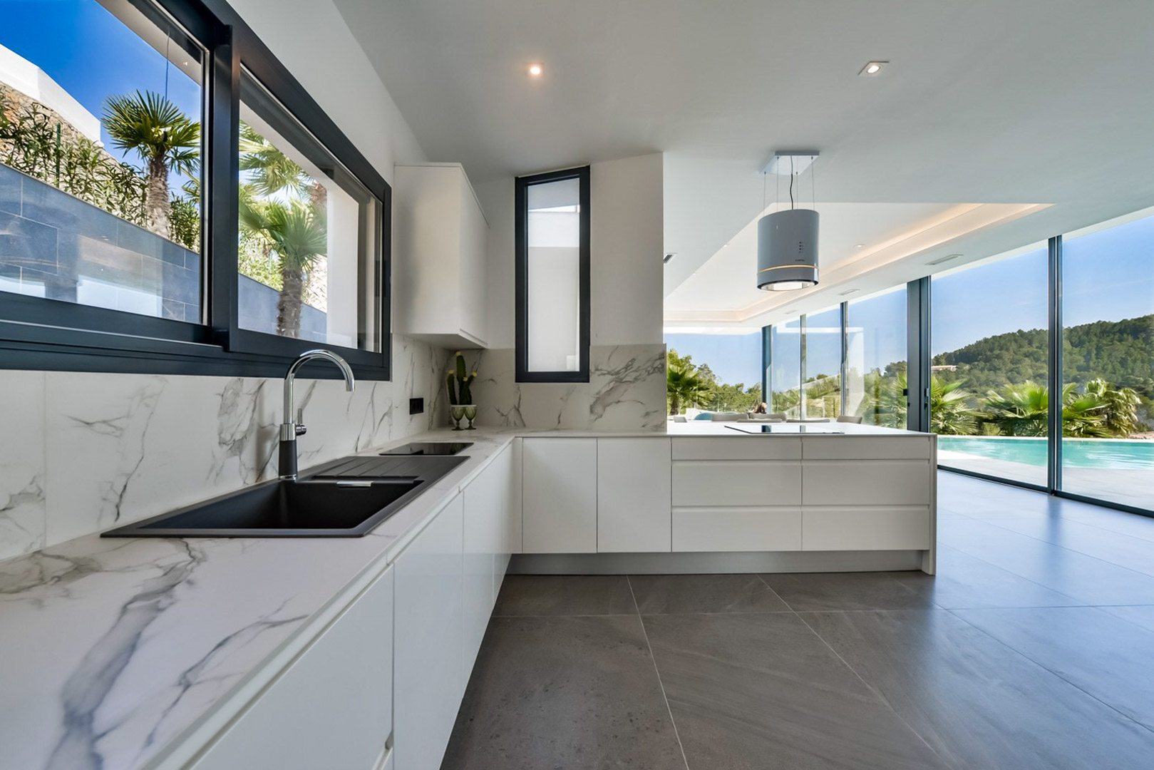 Cuisine contemporaine avec plans de travail en marbre et hotte cylindrique. Les grandes baies vitrées offrent une vue imprenable sur la piscine et les collines de Jávea.