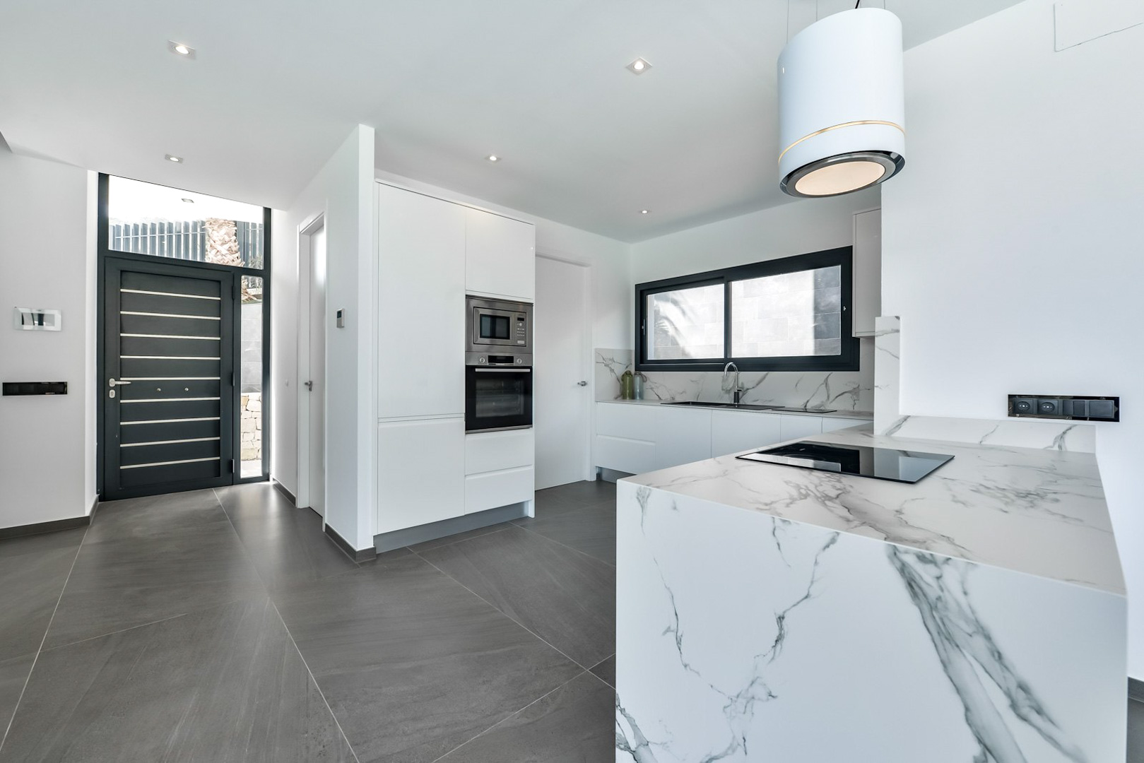 Cuisine moderne avec îlot en cascade effet marbre, électroménagers encastrés et hotte cylindrique, située près de l'entrée principale aux finitions noires.