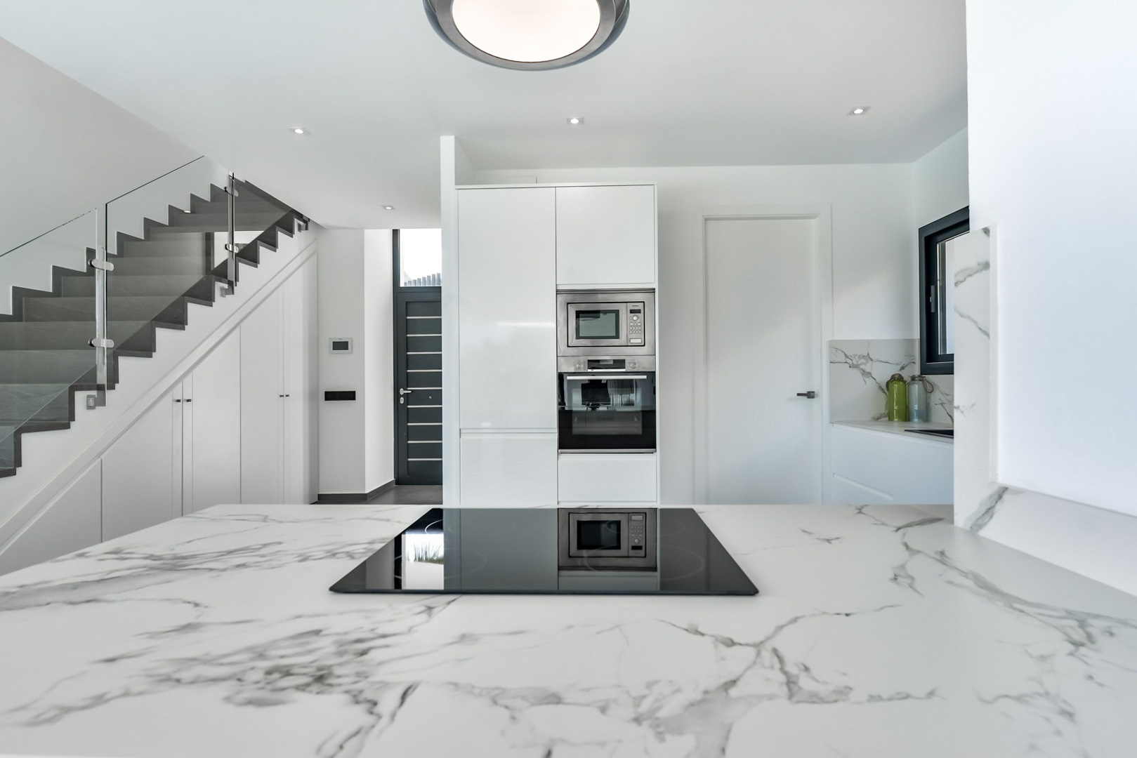Cuisine moderne avec plan de travail en marbre et plaque à induction, située à côté d'un escalier design avec garde-corps en verre et rangements intégrés.