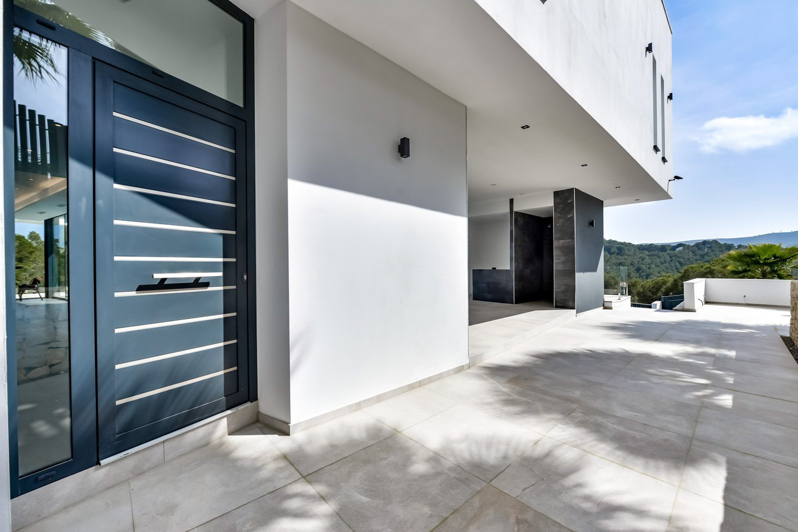 Entrée principale avec porte contemporaine bleu foncé à lattes métalliques, porche en grès cérame et vue dégagée sur les collines verdoyantes de Jávea.