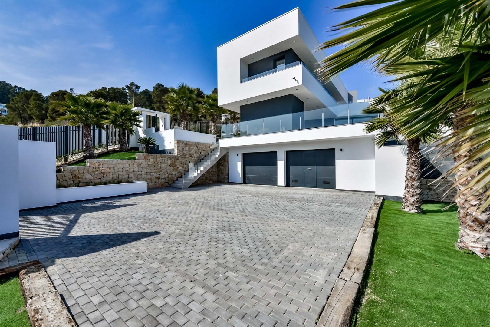 Façade contemporaine avec double garage, murs de soutènement en pierre naturelle et allée pavée entourée de végétation méditerranéenne.