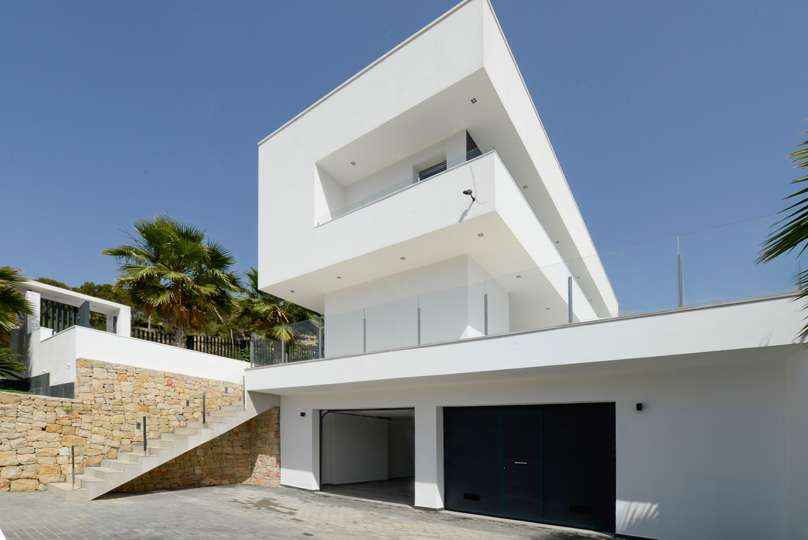 Façade moderne aux volumes blancs épurés, double garage, murs en pierre naturelle et escalier extérieur avec garde-corps en verre.