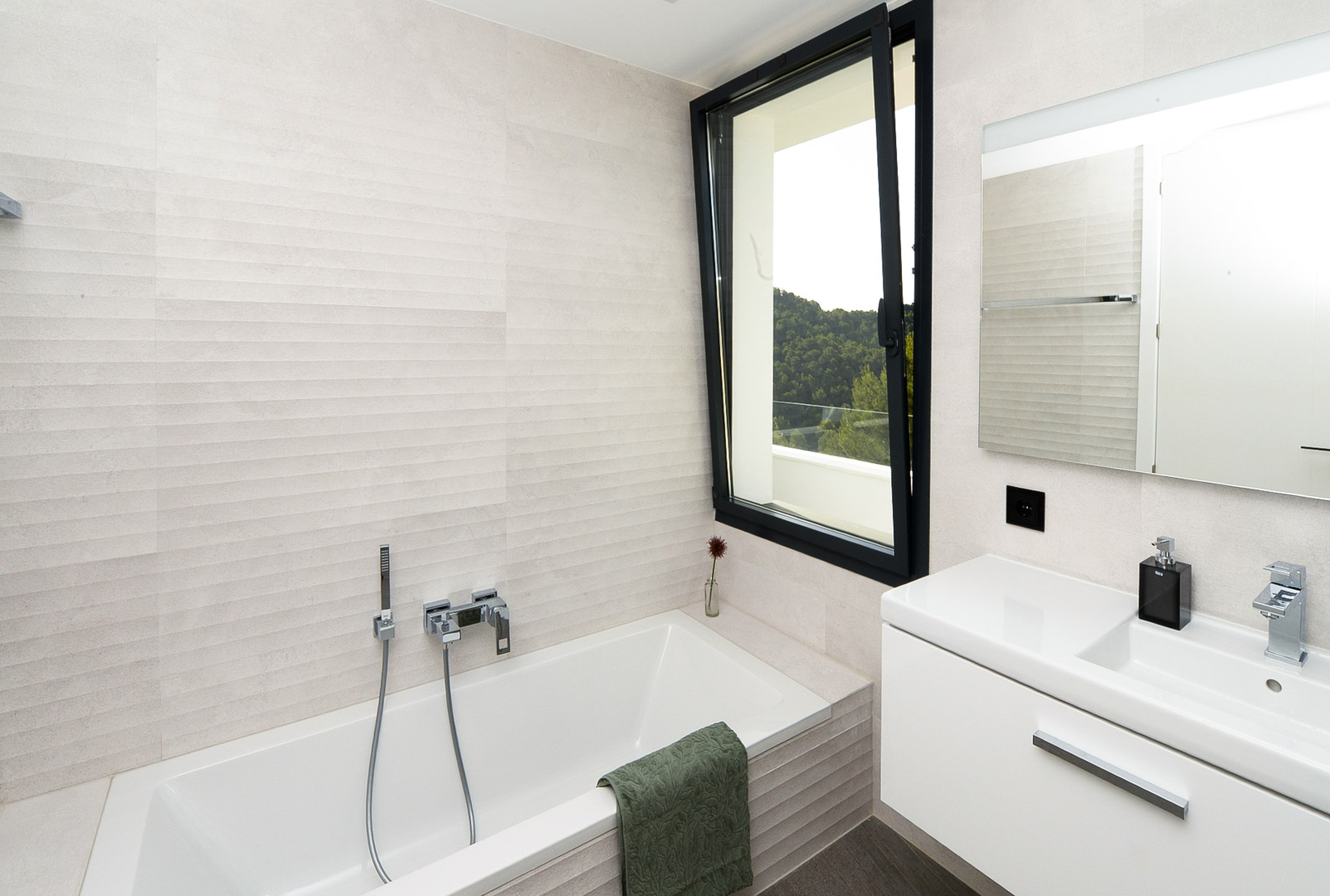 Salle de bain moderne avec baignoire encastrée, carrelage mural texturé et fenêtre ouvrant sur les collines verdoyantes de Jávea.