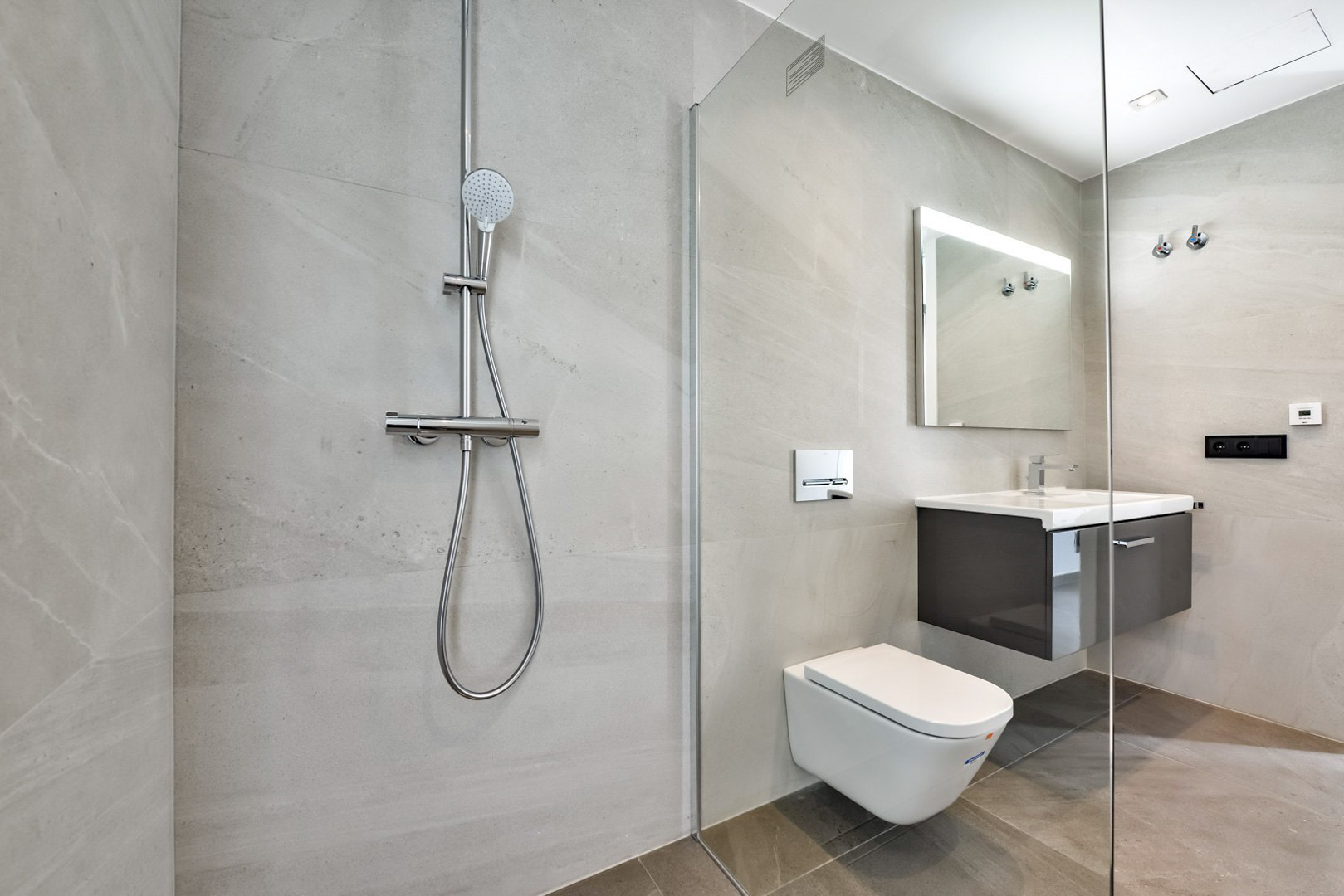 Salle de bain moderne avec douche à l'italienne, paroi vitrée, WC suspendu et meuble vasque flottant. Carrelage grand format gris et miroir rétroéclairé.