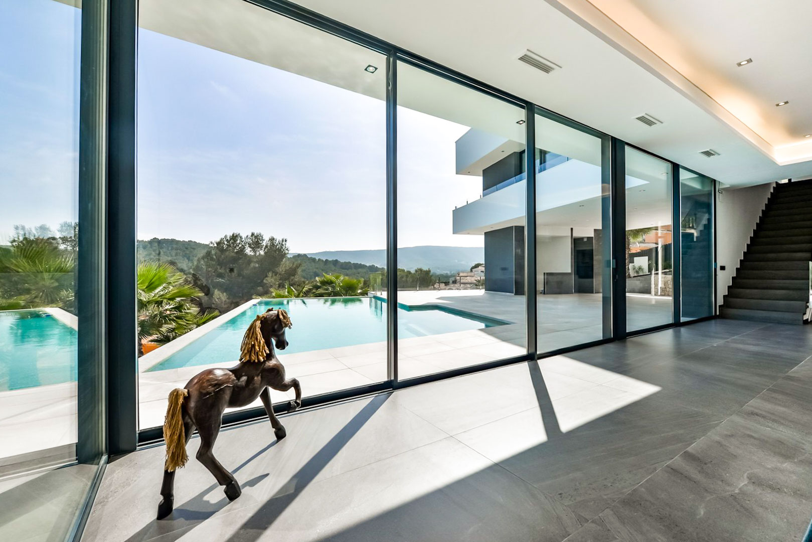 Salon avec baies vitrées toute hauteur offrant une vue sur la piscine à débordement. Sols en grès cérame gris et escalier contemporain intégré à l'espace de vie.