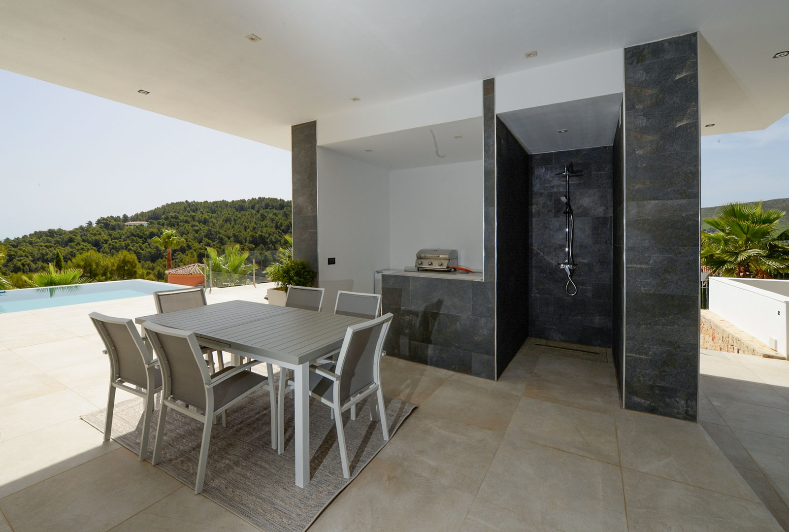 Terrasse couverte avec coin repas, cuisine d'été avec barbecue et douche extérieure en pierre sombre, offrant une vue sur la piscine et les collines.