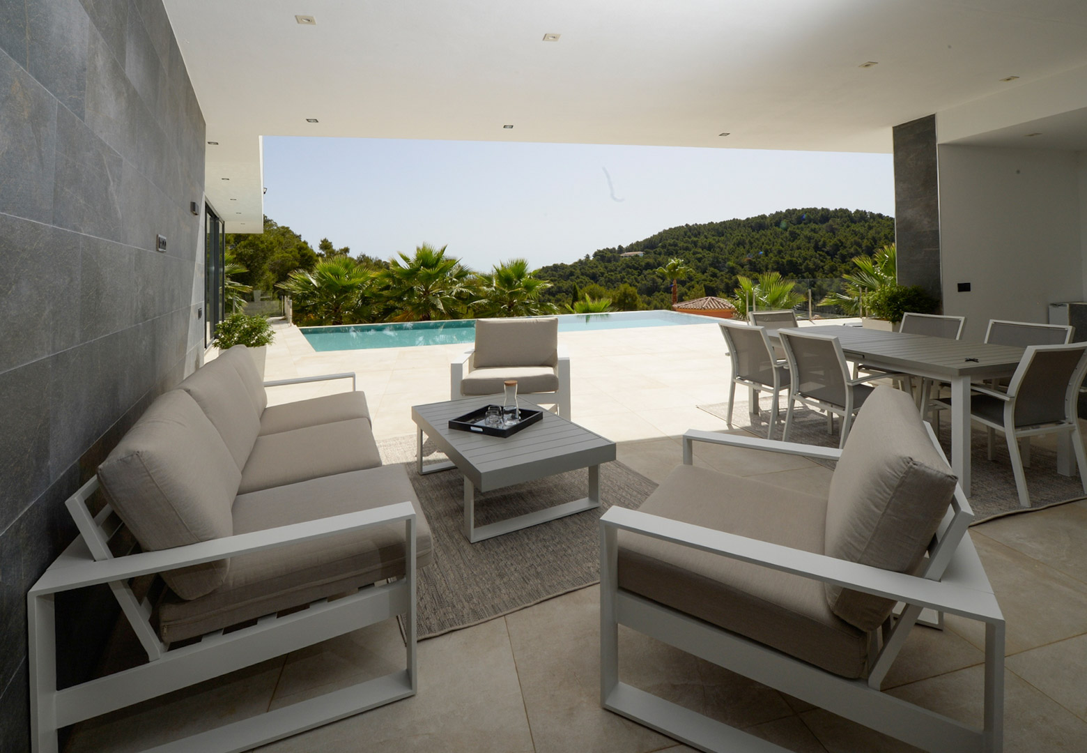Terrasse couverte avec salon et salle à manger d'été, offrant une vue dégagée sur la piscine à débordement et les collines verdoyantes.