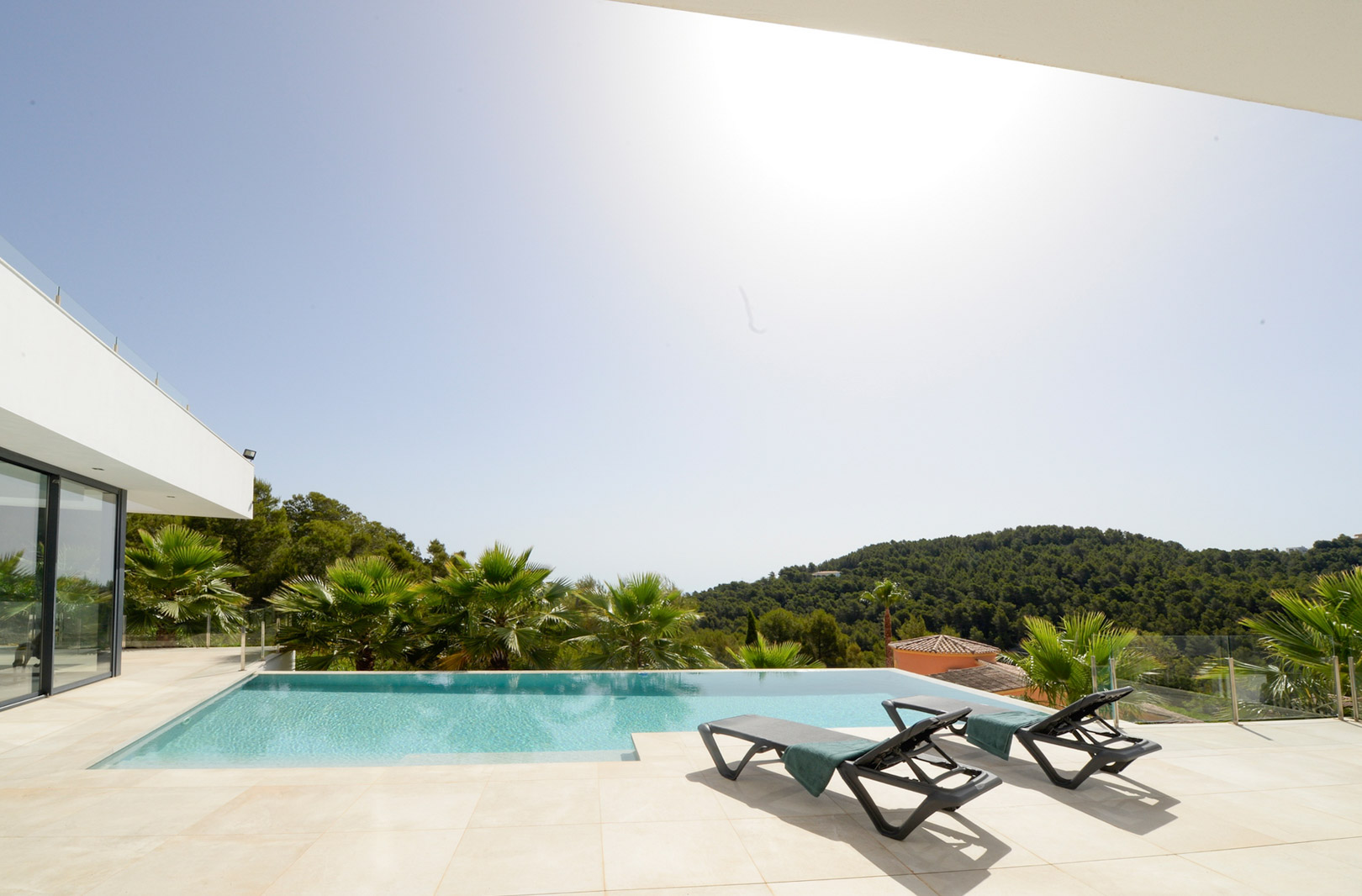 Terrasse ensoleillée avec piscine à débordement, chaises longues et vue panoramique sur les collines verdoyantes et la pinède environnante.
