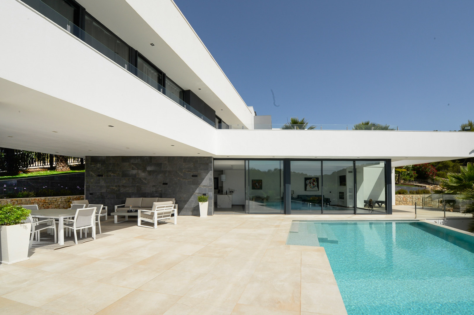 Terrasse principale avec piscine à débordement, salle à manger d'été couverte et architecture épurée aux larges baies vitrées et murs en pierre naturelle.