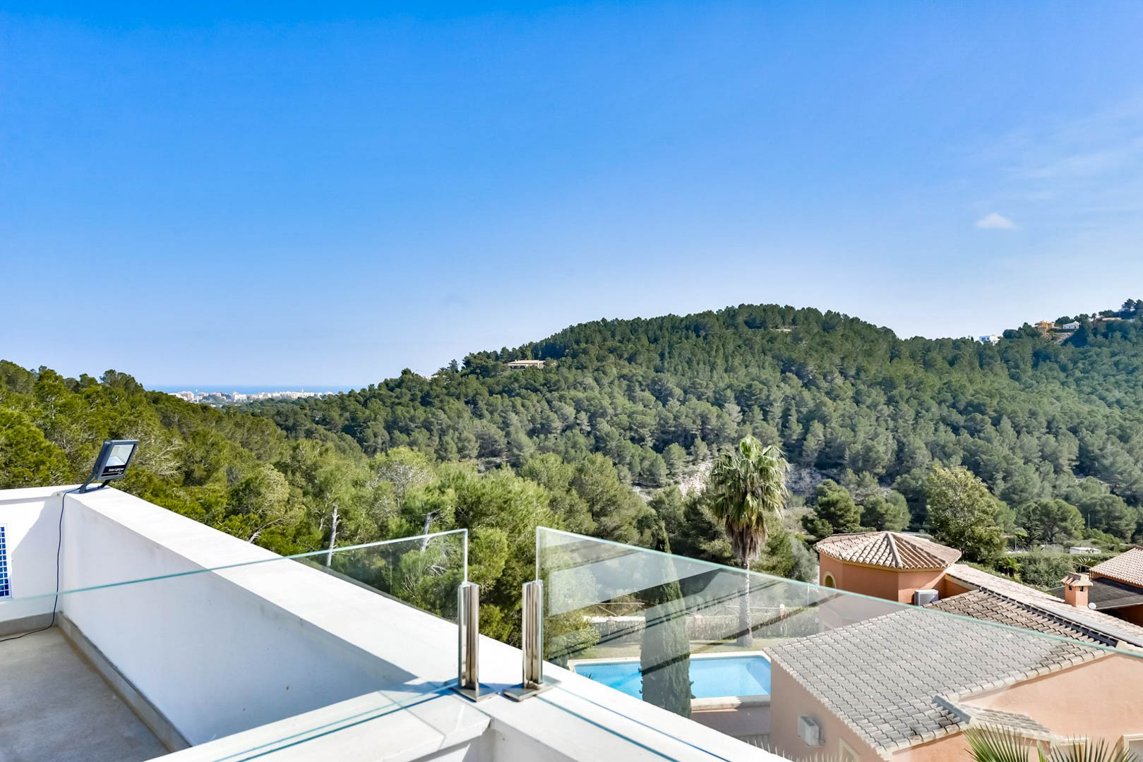Terrasse supérieure avec garde-corps en verre et acier inoxydable, offrant une vue panoramique sur les collines verdoyantes et la mer à l'horizon.