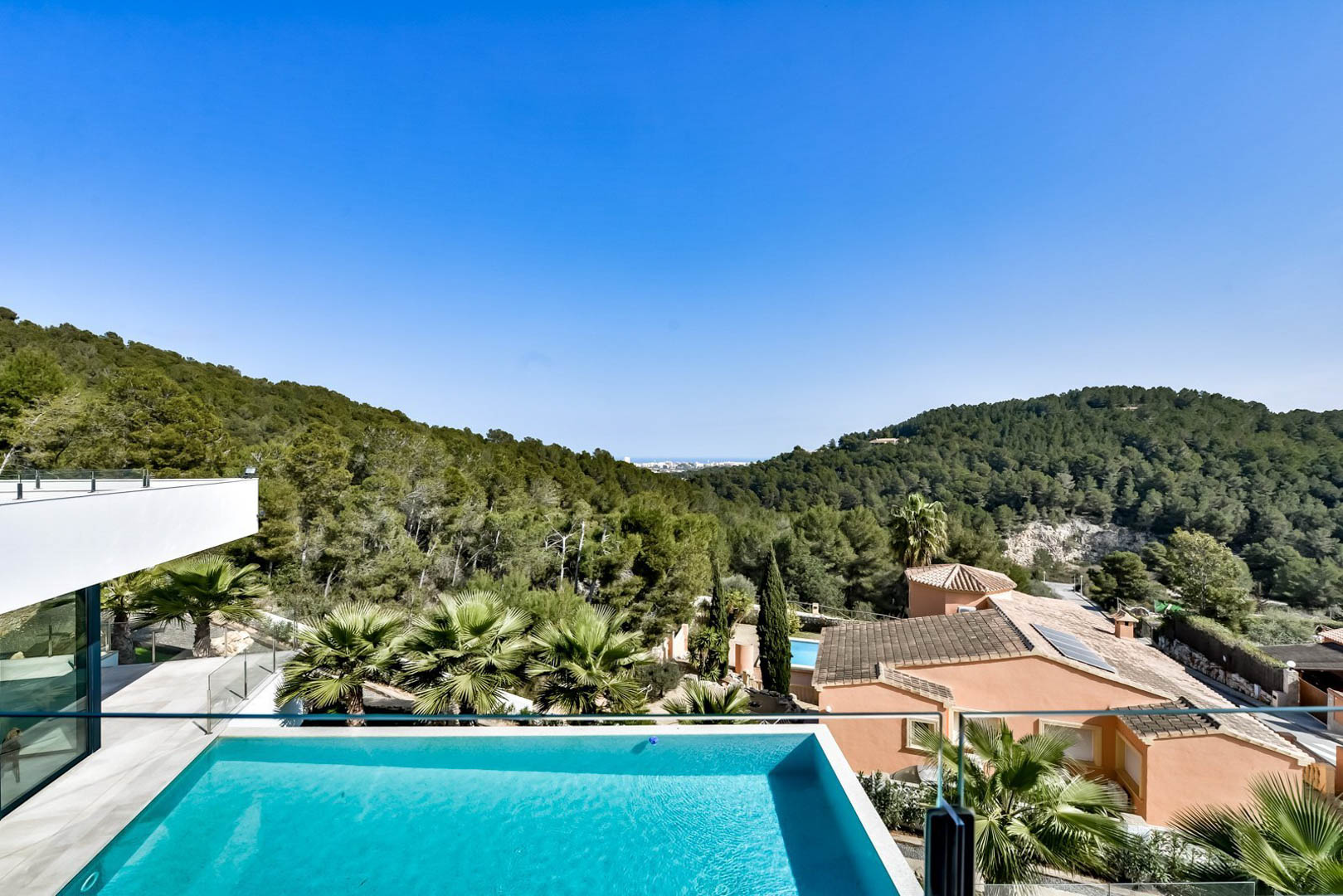 Vue panoramique depuis la terrasse supérieure avec piscine à débordement, garde-corps en verre et vue imprenable sur les collines verdoyantes et la mer.