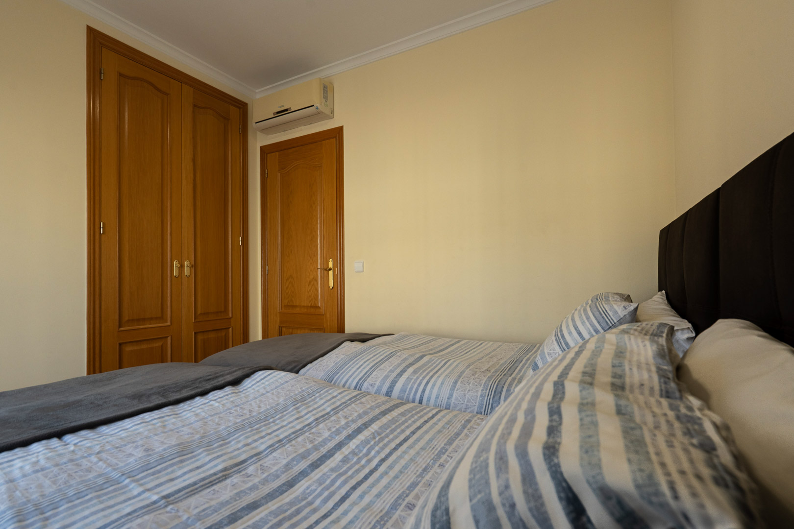 Chambre avec lits jumeaux, placards intégrés en bois et climatisation réversible. Murs de couleur crème et finitions soignées avec moulures au plafond.