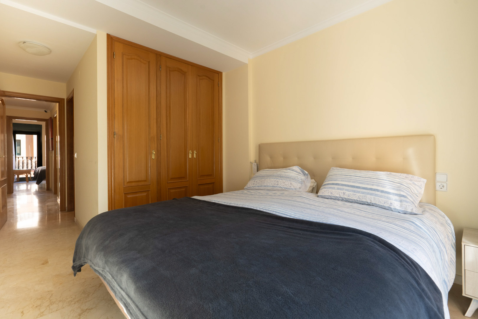 Chambre avec placards intégrés en bois, sol en marbre poli et tête de lit capitonnée, offrant un accès direct au couloir principal de l'appartement.