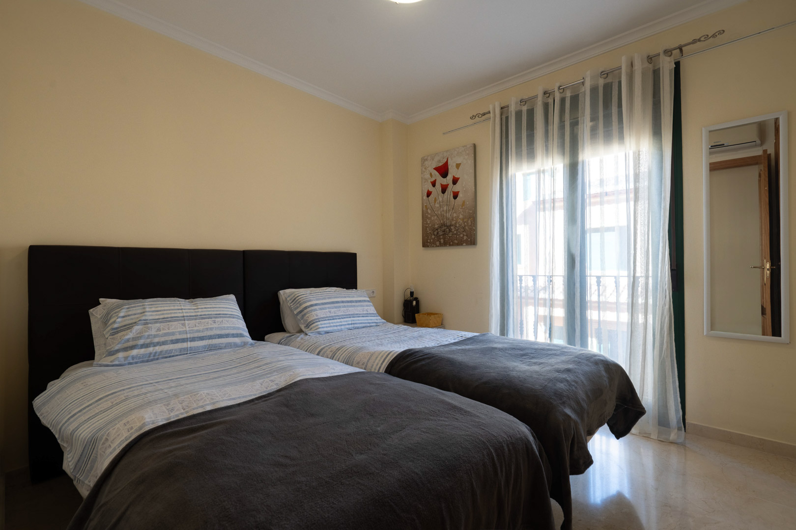 Chambre lumineuse avec deux lits simples, sol en marbre poli et accès au balcon protégé par des rideaux voilages.
