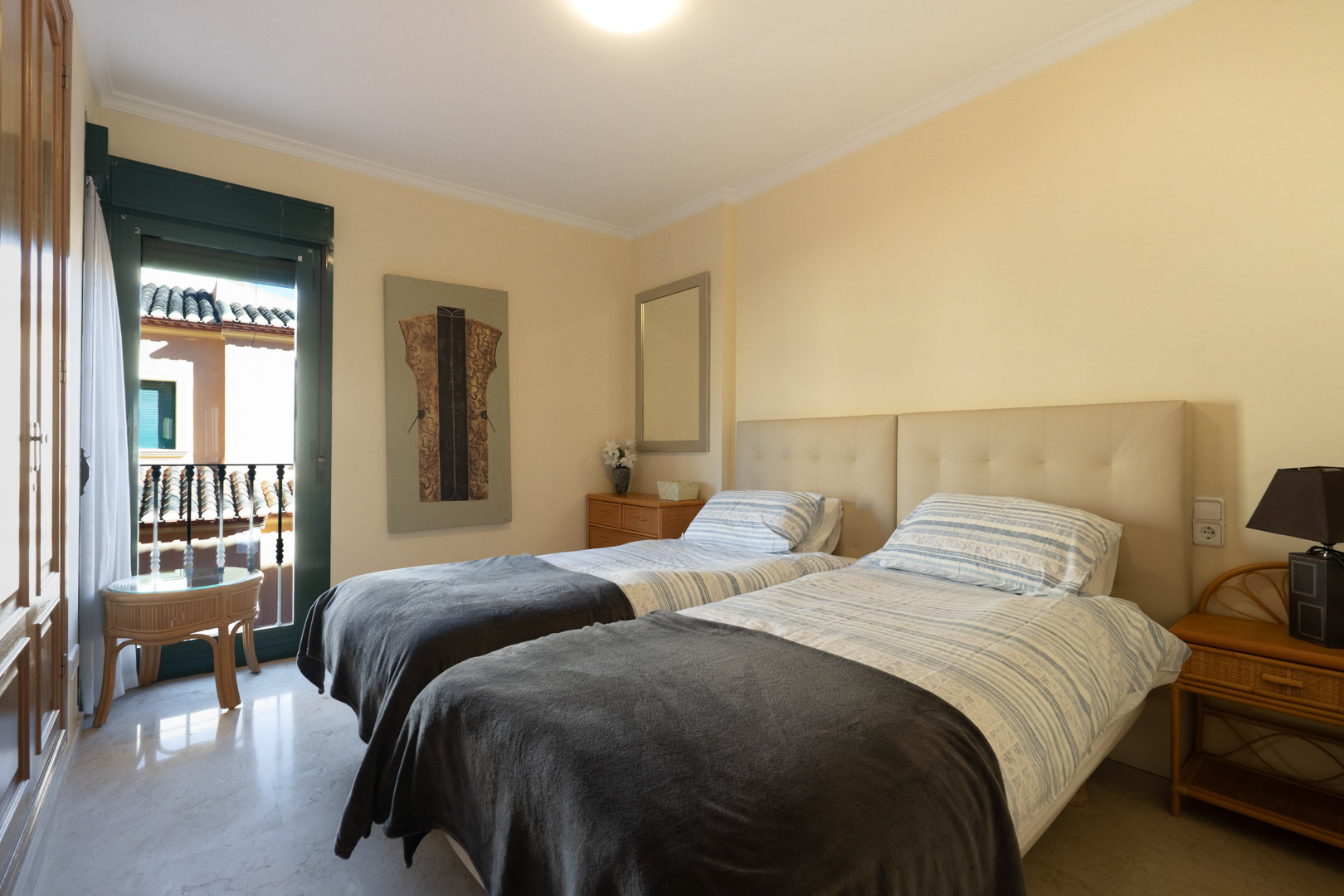 Chambre lumineuse avec deux lits simples, tête de lit rembourrée, sol en marbre et placards intégrés. Accès direct à un balcon privé.