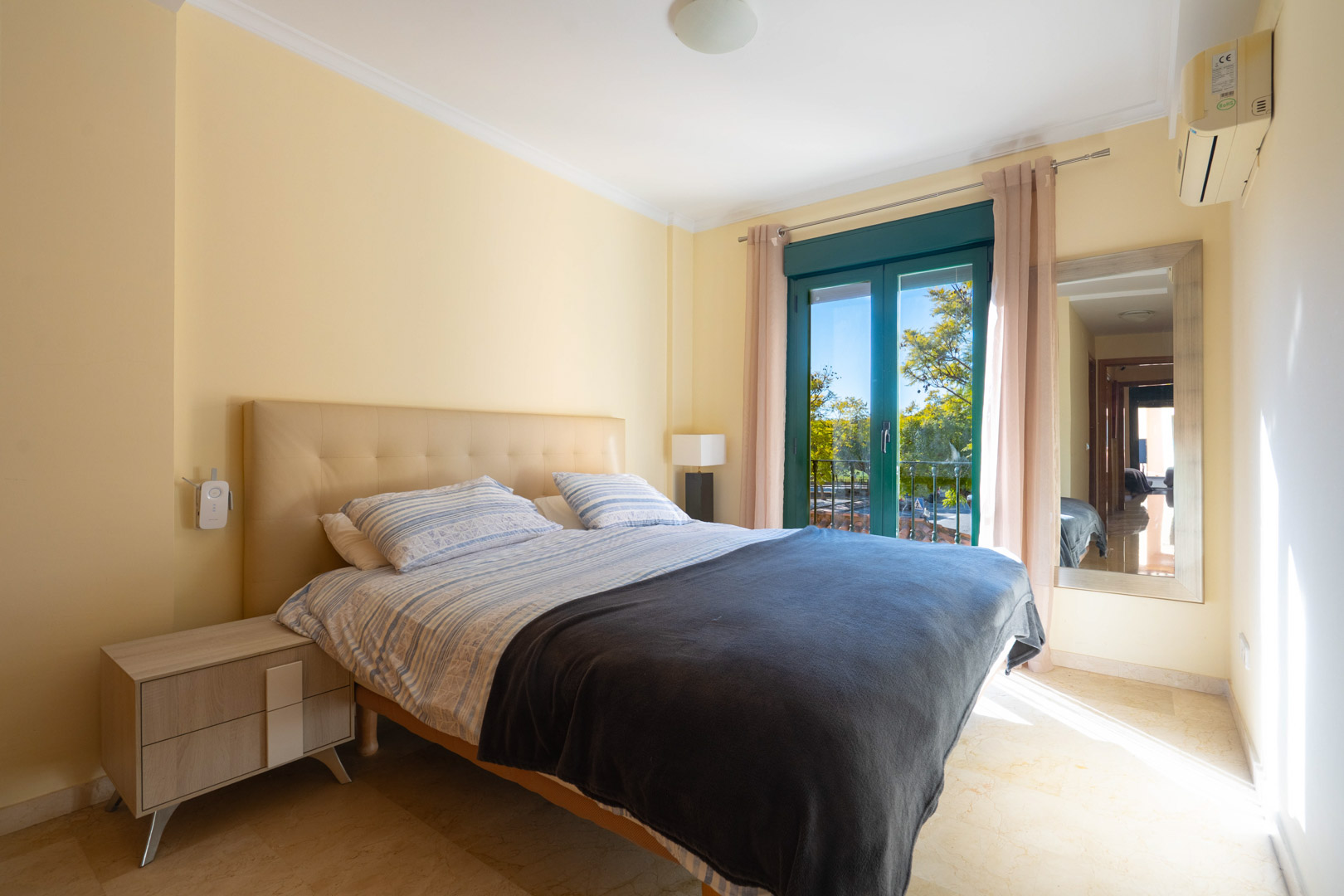 Chambre lumineuse avec sol en marbre, tête de lit capitonnée, climatisation et accès direct à un balcon donnant sur la végétation environnante.