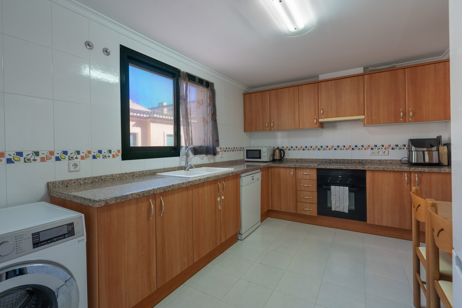 Cuisine fonctionnelle avec meubles en bois, plans de travail en granit, carrelage décoratif et équipement complet incluant lave-linge et lave-vaisselle.