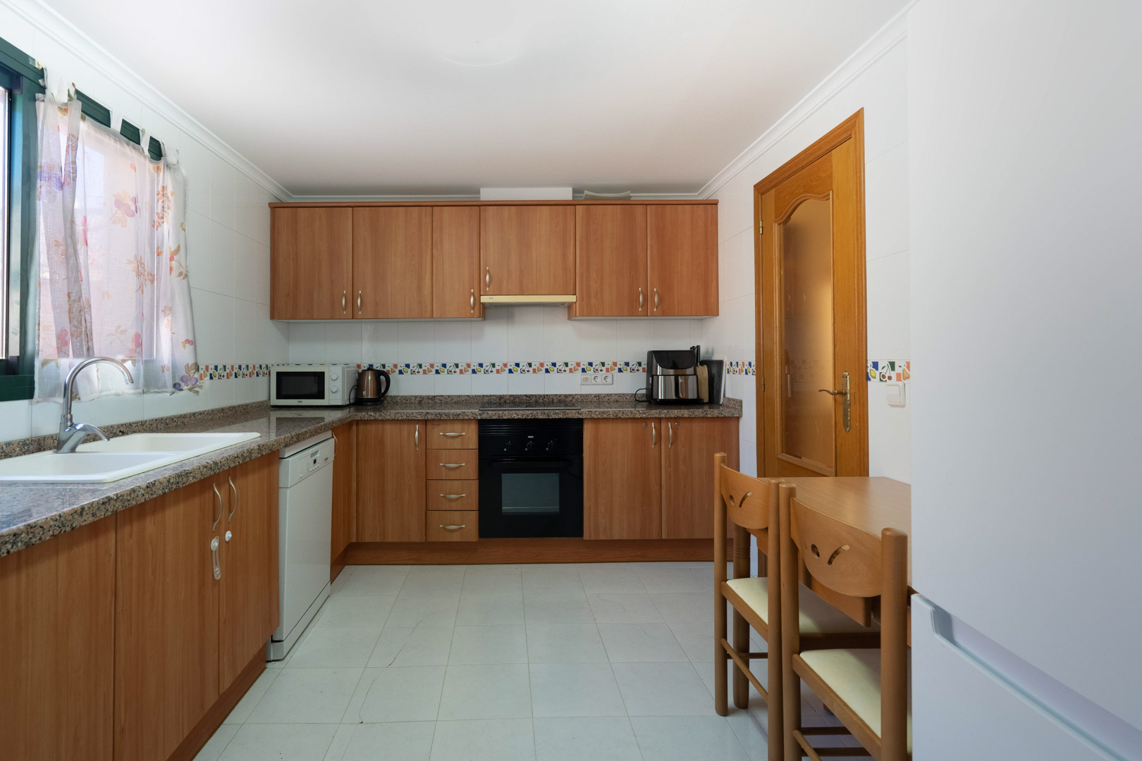 Cuisine indépendante avec placards en bois, plans de travail en granit et appareils intégrés. Elle comprend un coin repas compact et des murs carrelés avec frise décorative.