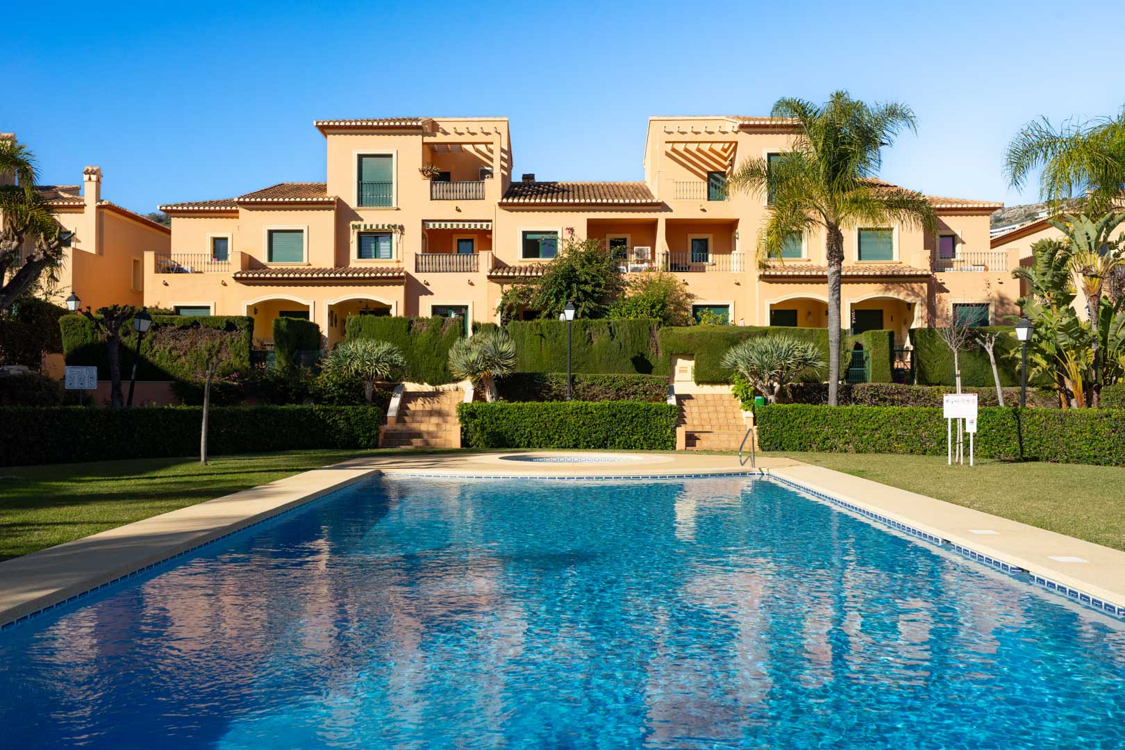 Piscine communautaire entourée de jardins méditerranéens et de pelouses, avec la façade traditionnelle du complexe Floridamar en arrière-plan sous un ciel bleu.