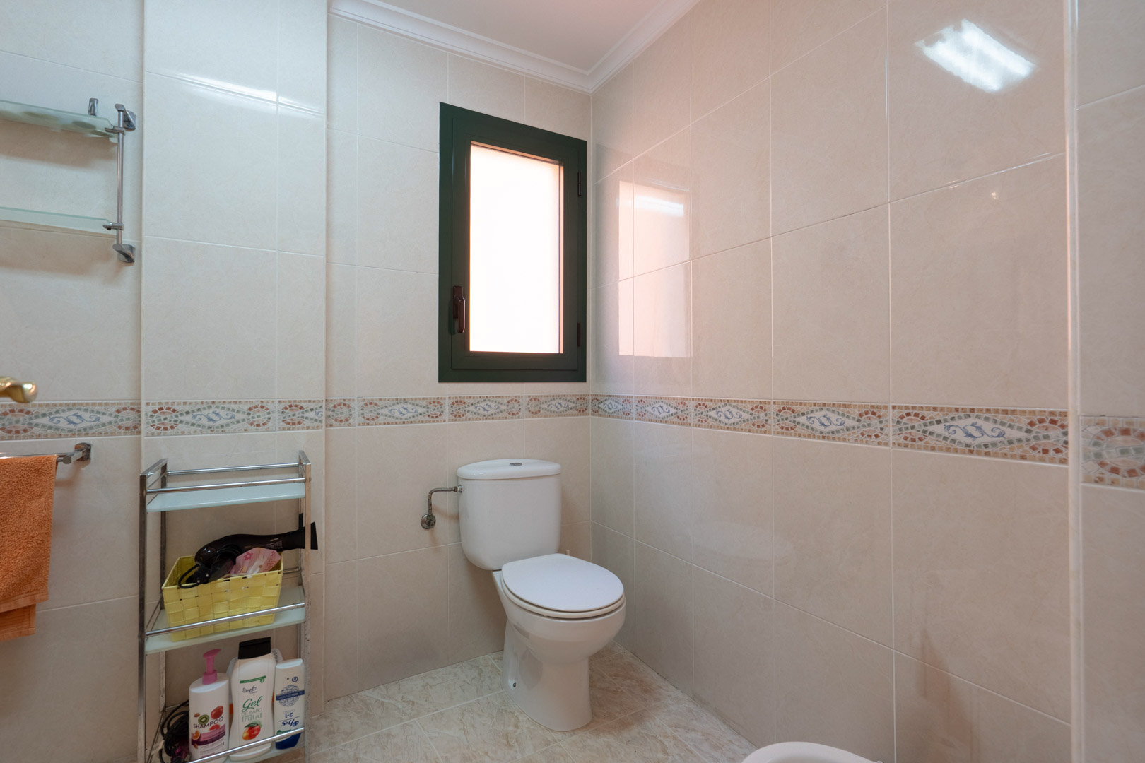 Salle de bains avec carrelage crème, frise décorative en mosaïque, toilettes blanches et une fenêtre oscillo-battante assurant luminosité et aération.
