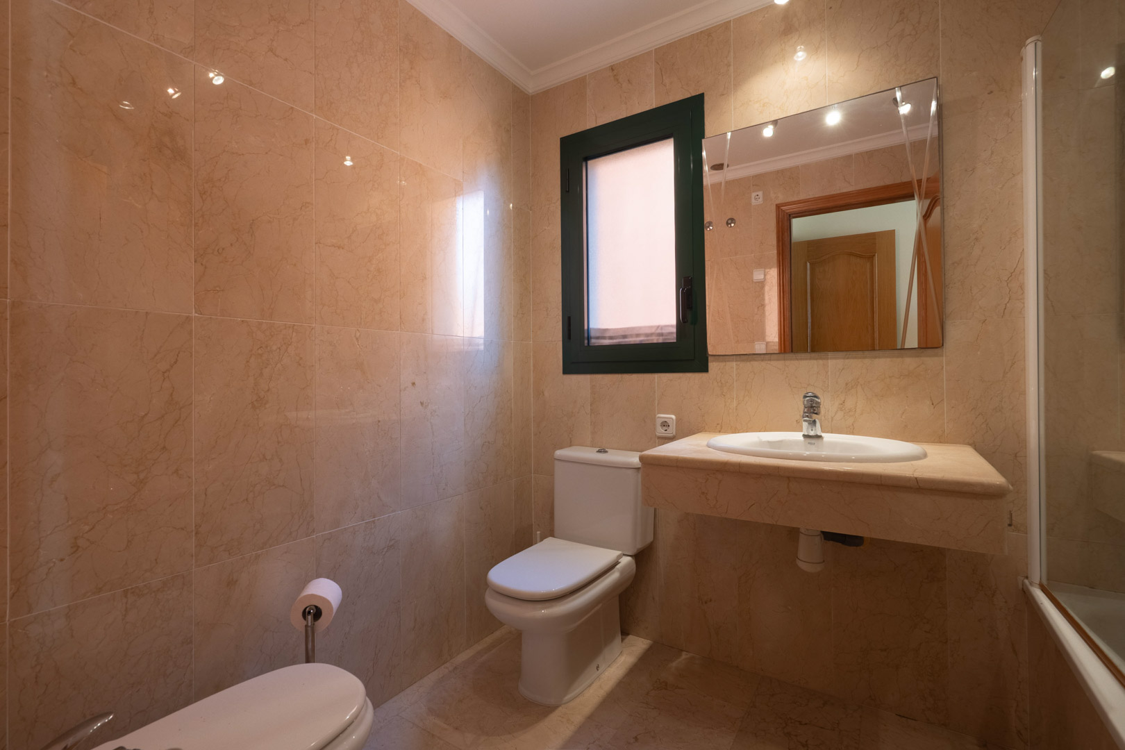 Salle de bains revêtue de marbre crème, comprenant un plan de toilette en pierre avec vasque intégrée, des sanitaires blancs et une fenêtre d'aération.
