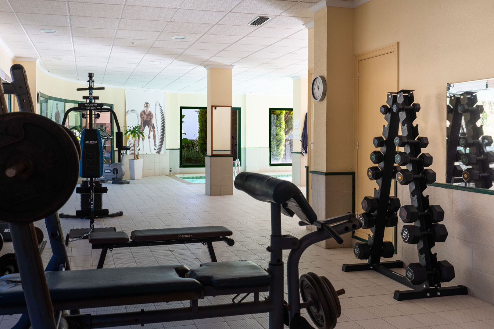 Salle de sport pour résidents équipée de machines de musculation et d'haltères, située à côté de la piscine intérieure avec vue sur le jardin.