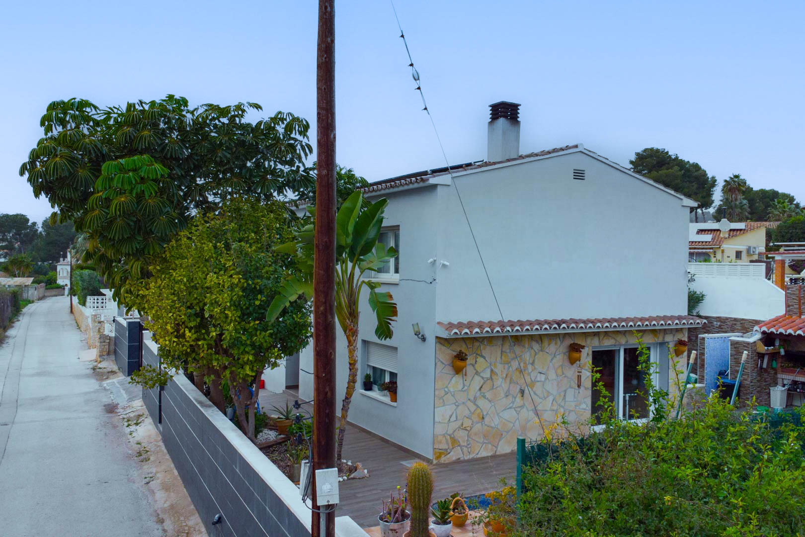 Vue extérieure de la villa présentant une façade blanche avec parement en pierre naturelle, entourée de végétation méditerranéenne et de panneaux solaires.