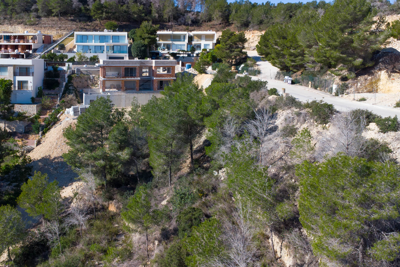Terrain à bâtir surélevé à Tosalet 5, entouré de pins et de villas contemporaines. On aperçoit la route d'accès goudronnée et une structure en cours de construction.