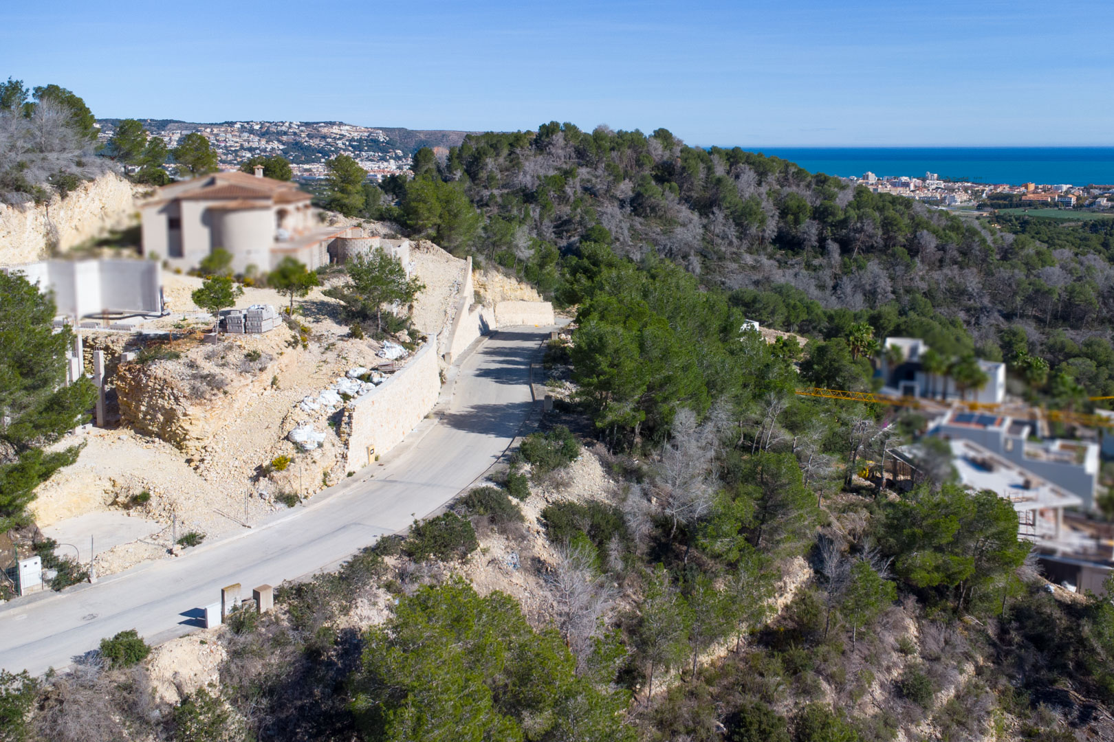 Vue aérienne de la route d'accès asphaltée à Tosalet 5, bordée de murs de soutènement en pierre et de pins, offrant une perspective sur la mer et la côte de Jávea.