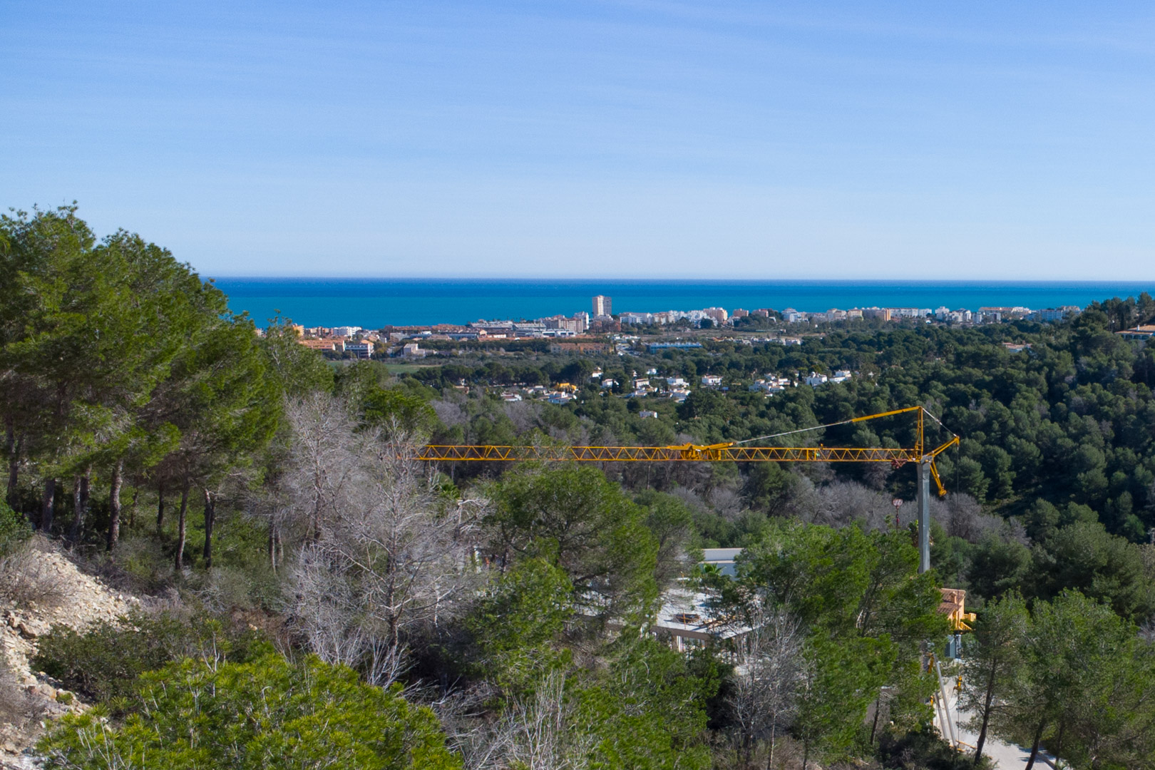 Vue panoramique sur la mer Méditerranée et le littoral de Jávea depuis le terrain, entouré d'une pinède dense sous un ciel bleu azur.