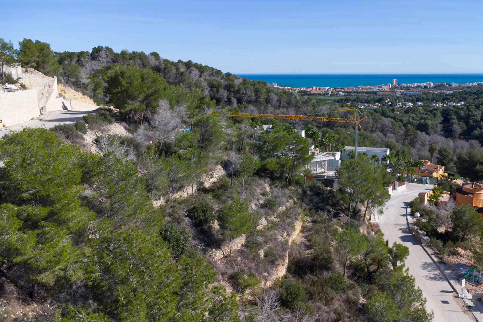 Vue surélevée d'un terrain en pente avec pins méditerranéens, murs de soutènement en pierre et vue panoramique sur la mer et la ville de Jávea.