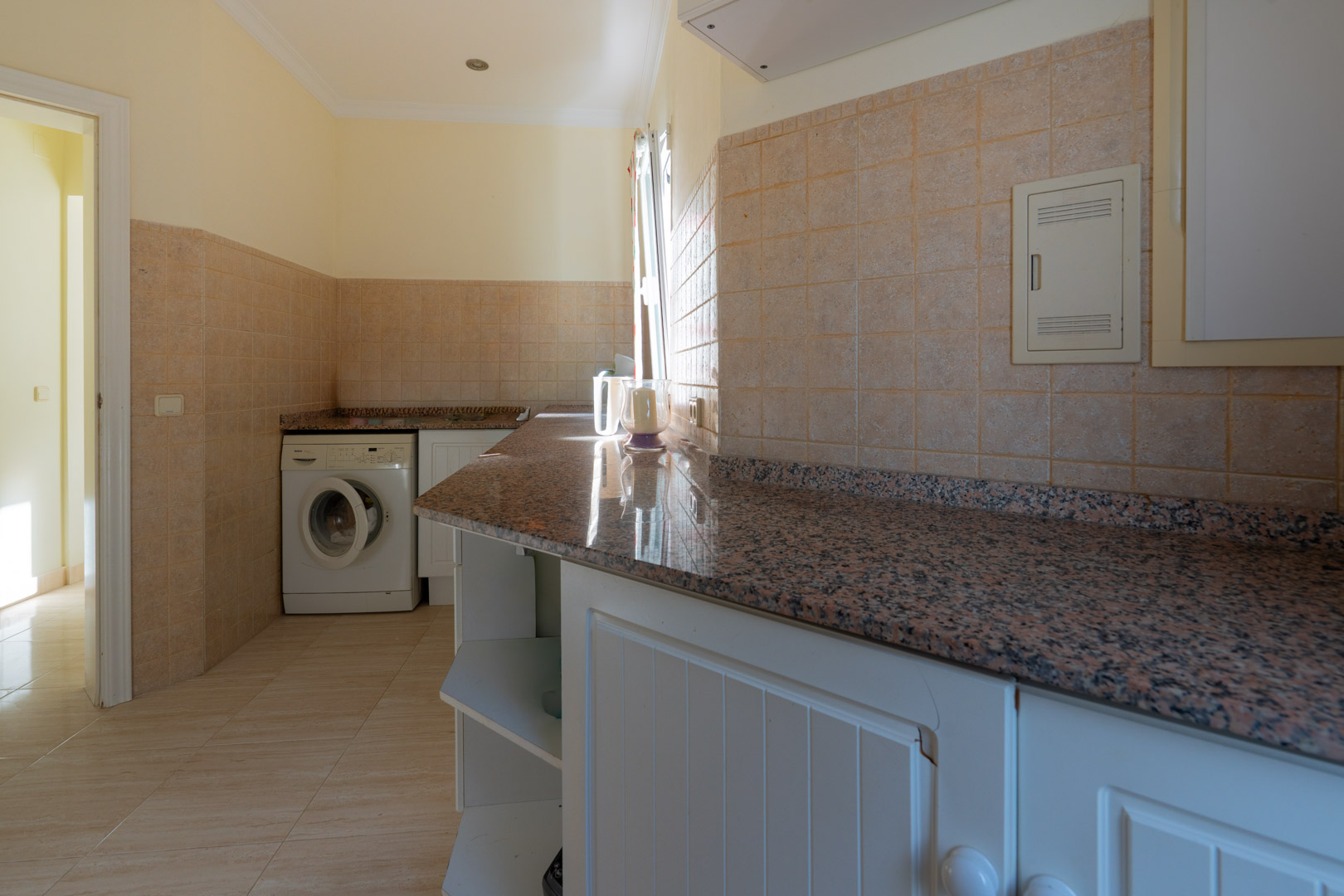 Buanderie pratique avec plans de travail en granit, meubles blancs et lave-linge, dotée de carrelage en céramique et d'un éclairage naturel.