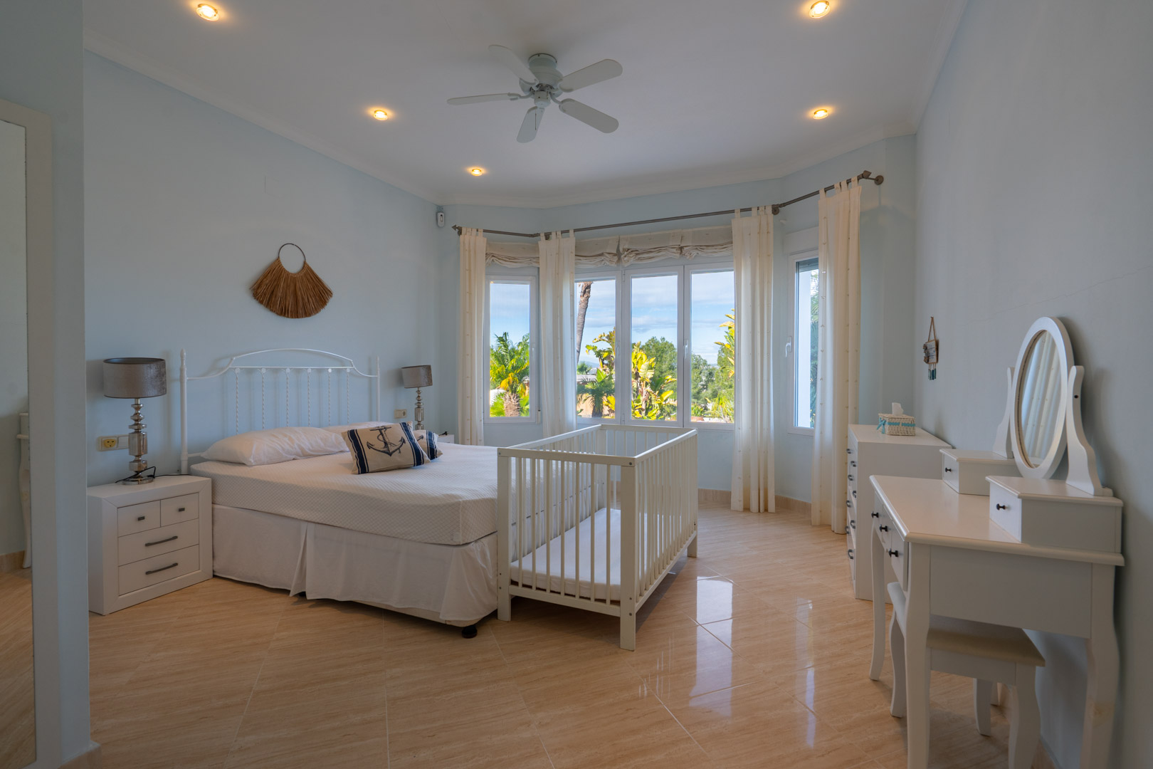 Chambre avec sol en grès poli, baie vitrée donnant sur la végétation, mobilier blanc, coiffeuse et ventilateur de plafond.